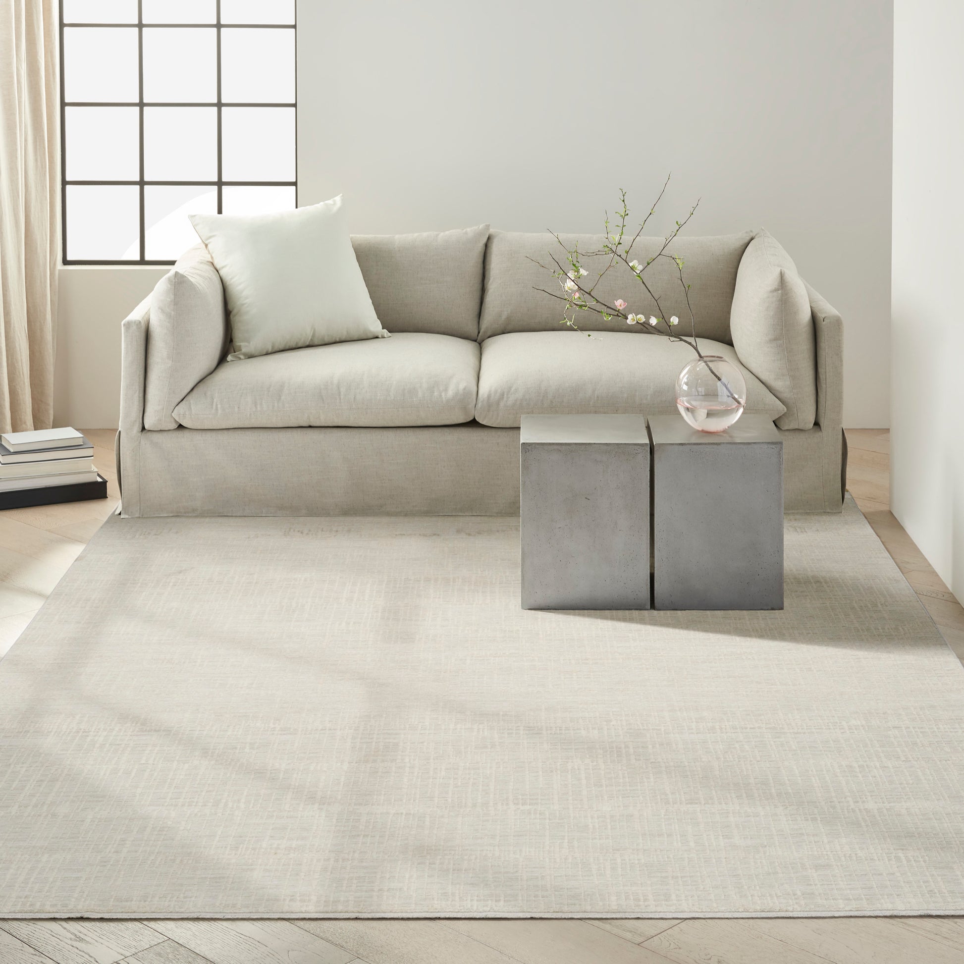 Light gray sofa with plush cushions, concrete block side tables, glass vase with branches, beige rug, minimalist decor.