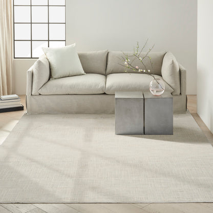 Light gray sofa with plush cushions, concrete block side tables, glass vase with branches, beige rug, minimalist decor.