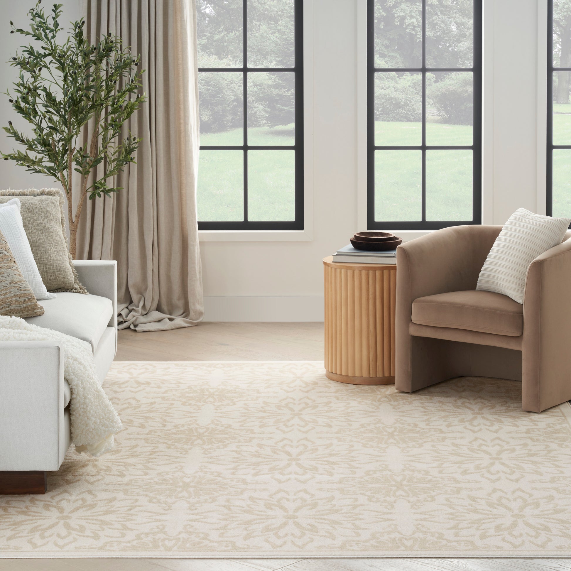 Neutral living room with beige armchair, white sofa, cream damask rug, round wood side table, plant, black-framed windows.