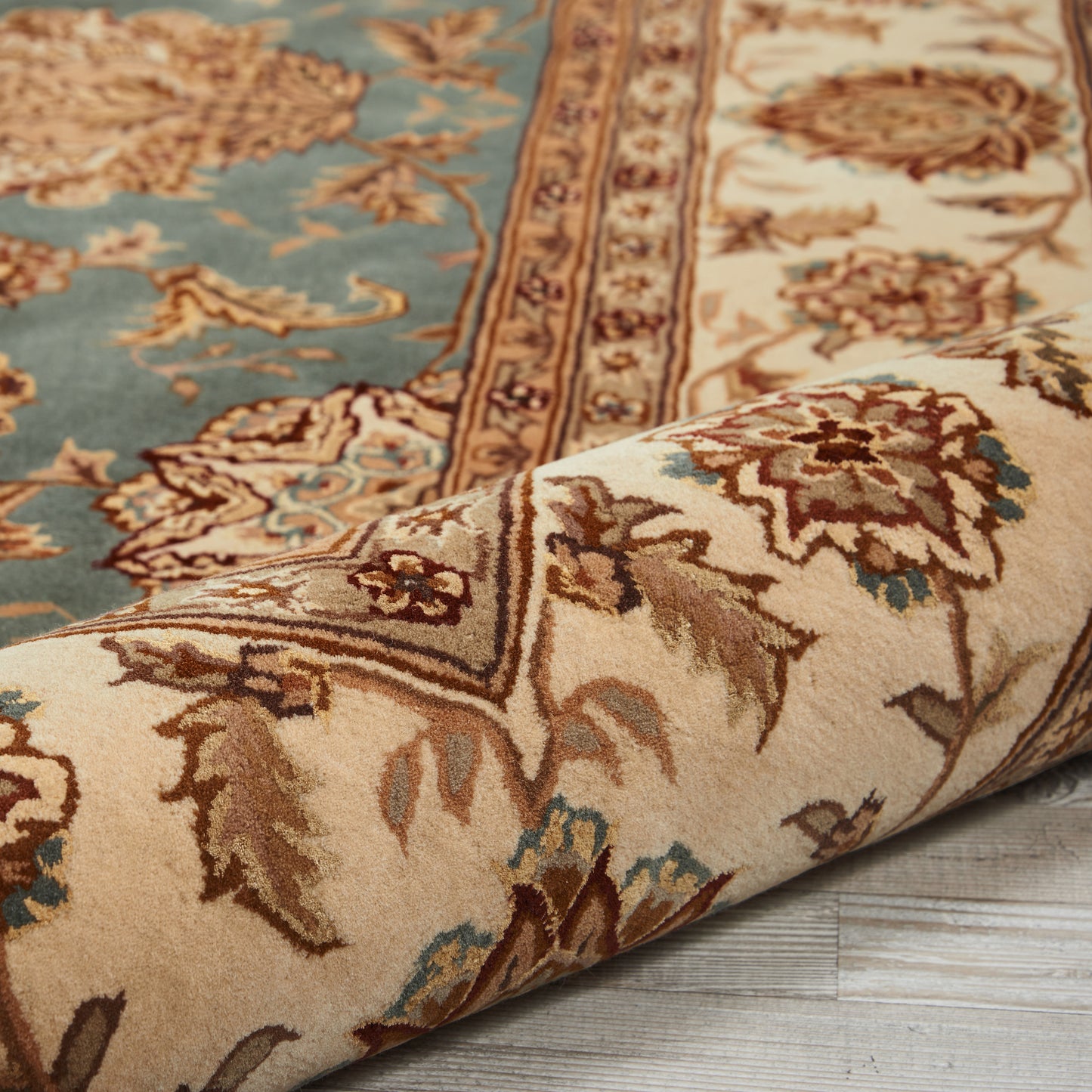 Traditional Persian-style rug in blue, beige and brown with ornate floral border; rolled corner on a wood floor.