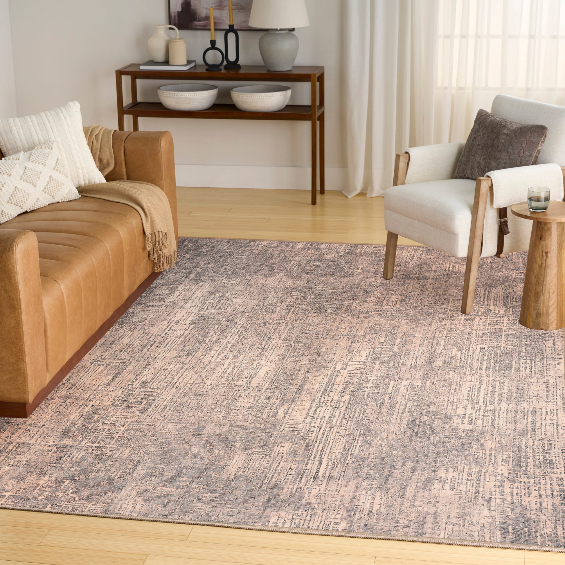 Cozy living room with tan leather sofa, gray patterned rug, white armchair, and wooden side table near sheer curtains.