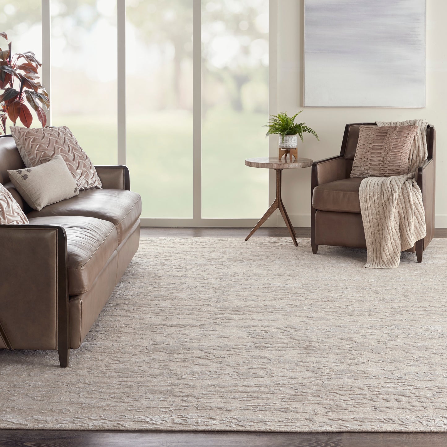 Taupe leather sofa with matching armchair, beige pillows and throw, round wood side table with fern, neutral rug.