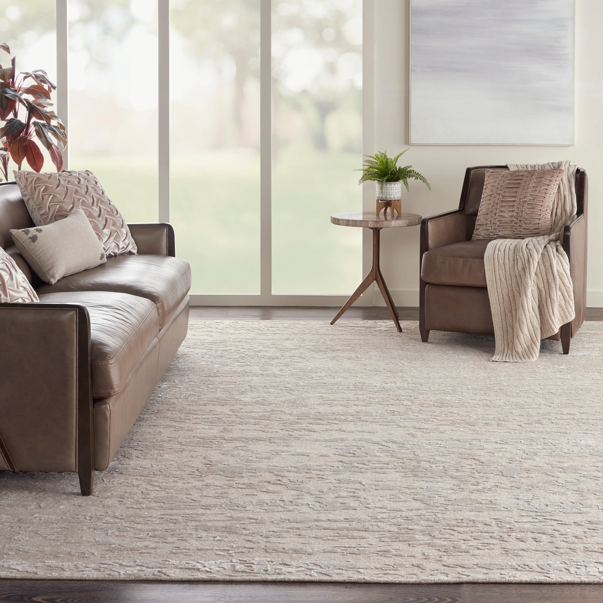 Taupe leather sofa with matching armchair, beige pillows and throw, round wood side table with fern, neutral rug.