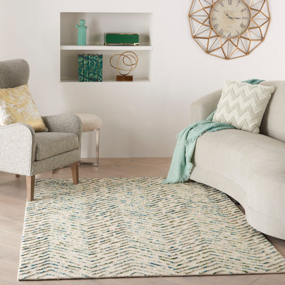 Beige curved sofa with mint throw & chevron pillow, gray armchair, blue-green patterned rug, white shelves, gold clock.