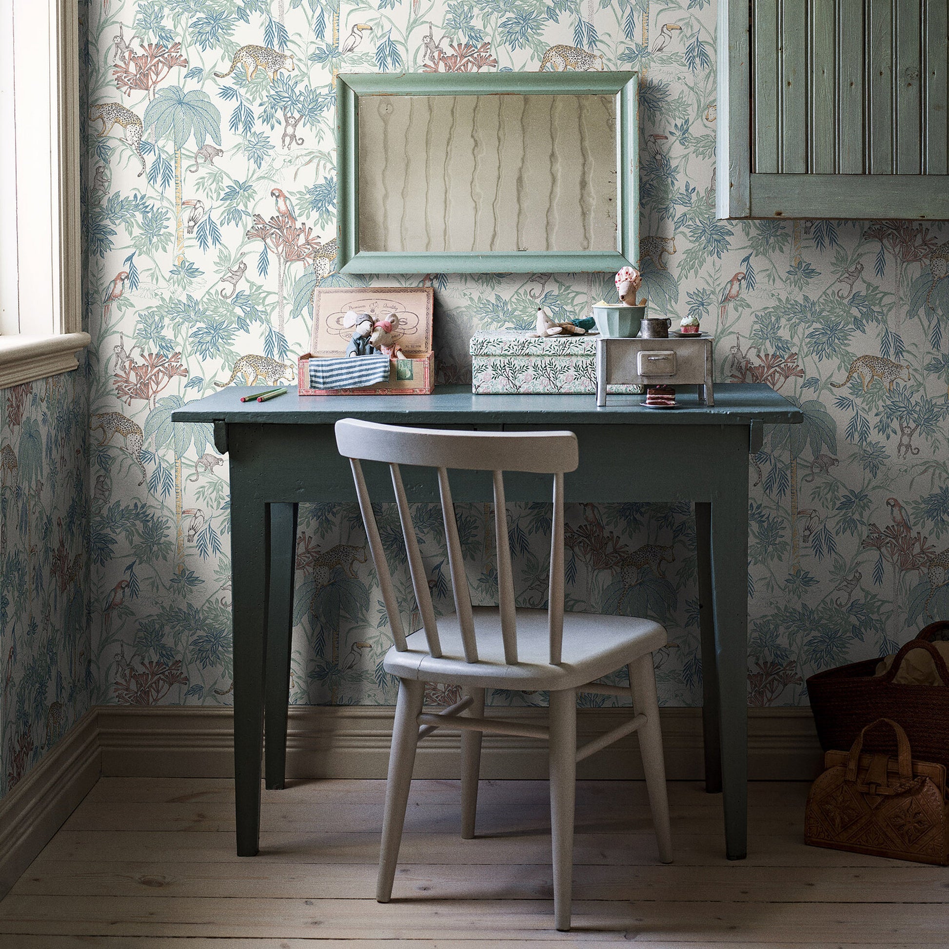 Dark teal wooden desk with straight legs, light gray chair, vintage floral wallpaper; mini mirror above, desk accessories.