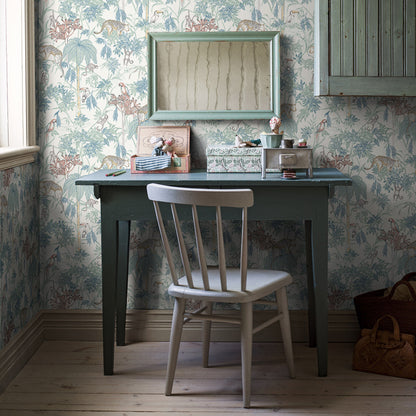 Dark teal wooden desk with straight legs, light gray chair, vintage floral wallpaper; mini mirror above, desk accessories.