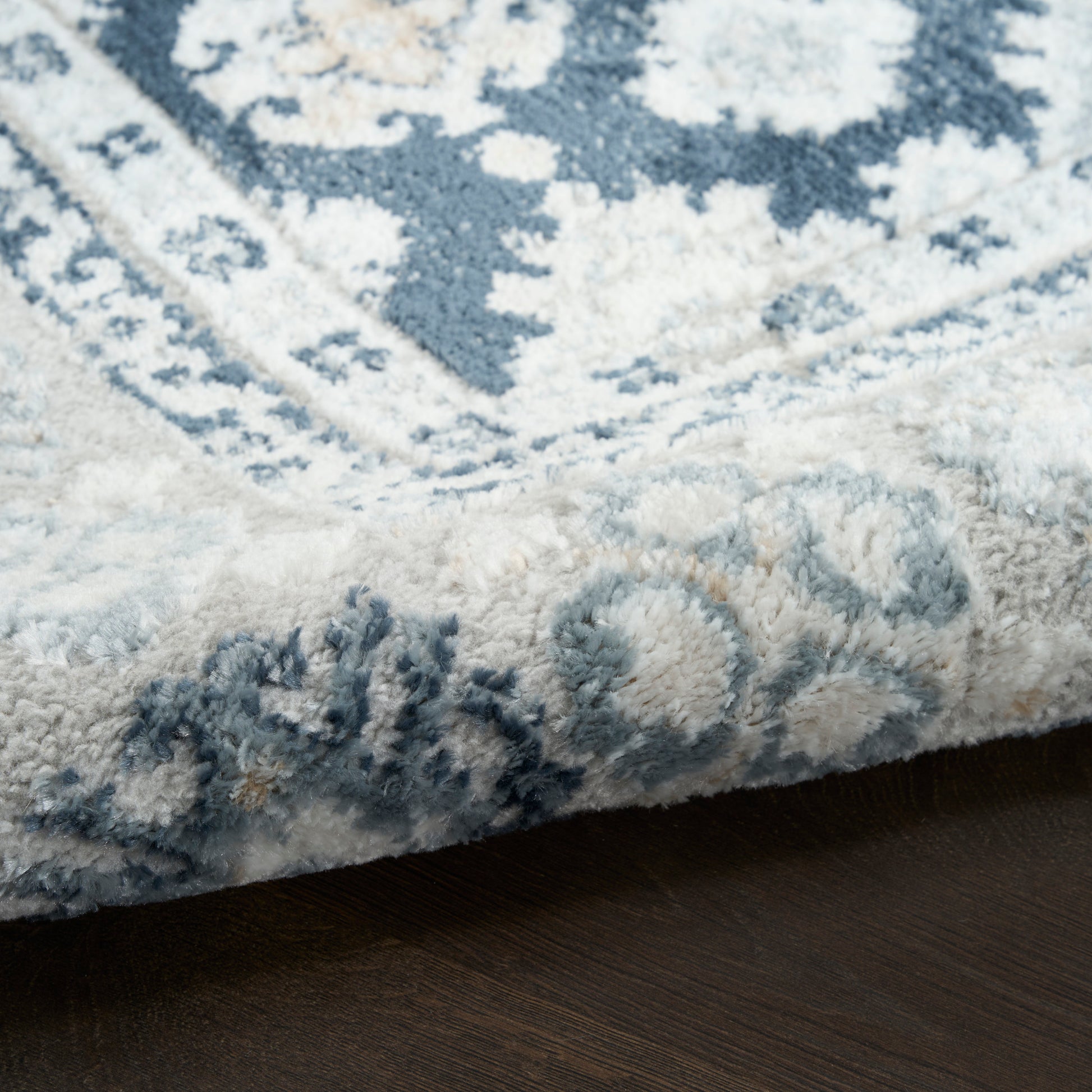 Blue and ivory ornate patterned plush rug with thick pile, edge folded on dark wood floor.