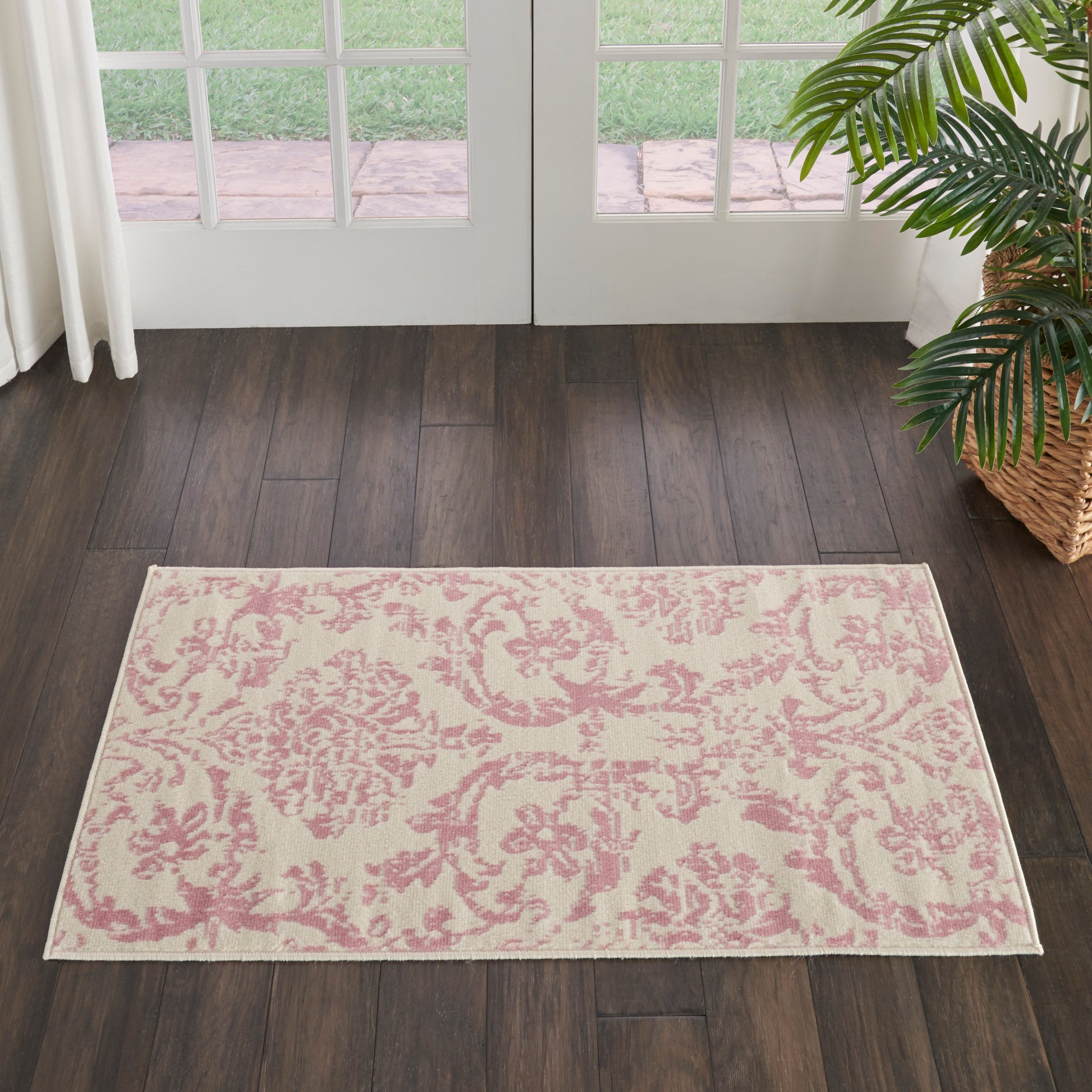 Rectangular cream area rug with pink damask floral pattern; placed on dark wood floor by glass French doors.