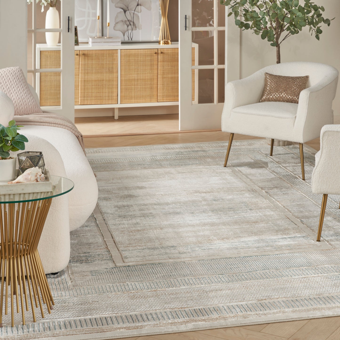Bright living room with cream armchairs, light gray patterned rug, round glass side table on a gold base, and greenery.