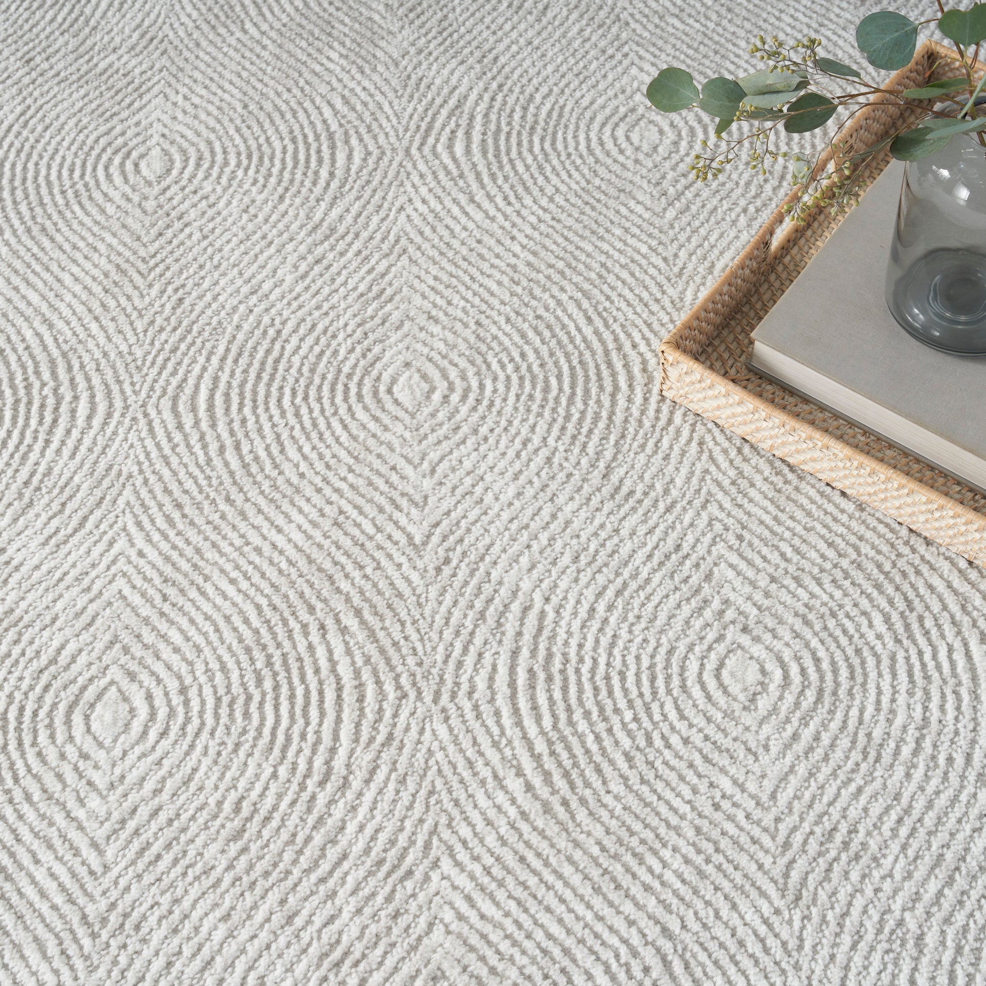 Light gray textured rug featuring concentric loop pattern; wicker tray with books and a glass vase of eucalyptus.