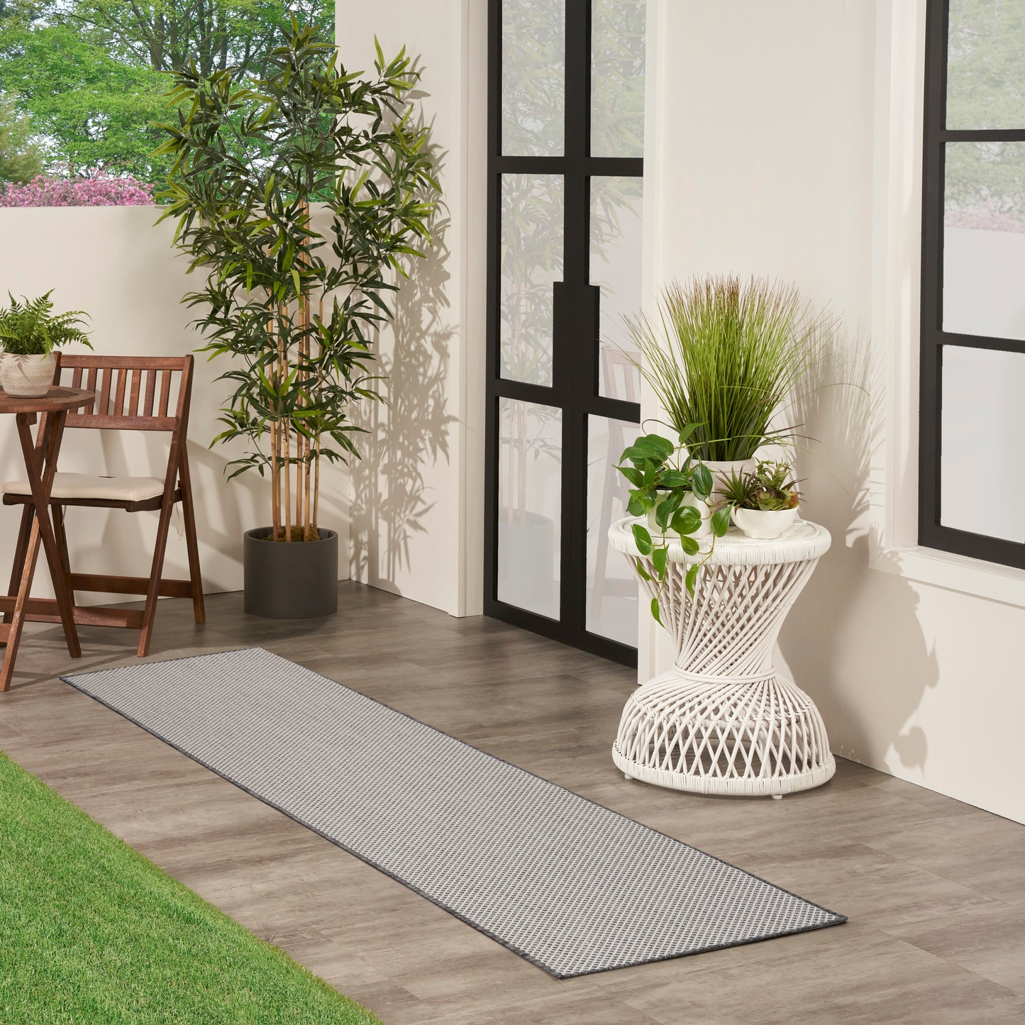 Modern sunlit patio with black-framed glass doors, gray striped rug, tall potted plant, and wicker side table with plants.