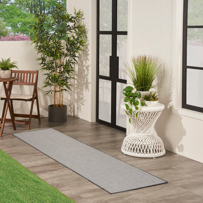 Modern sunlit patio with black-framed glass doors, gray striped rug, tall potted plant, and wicker side table with plants.