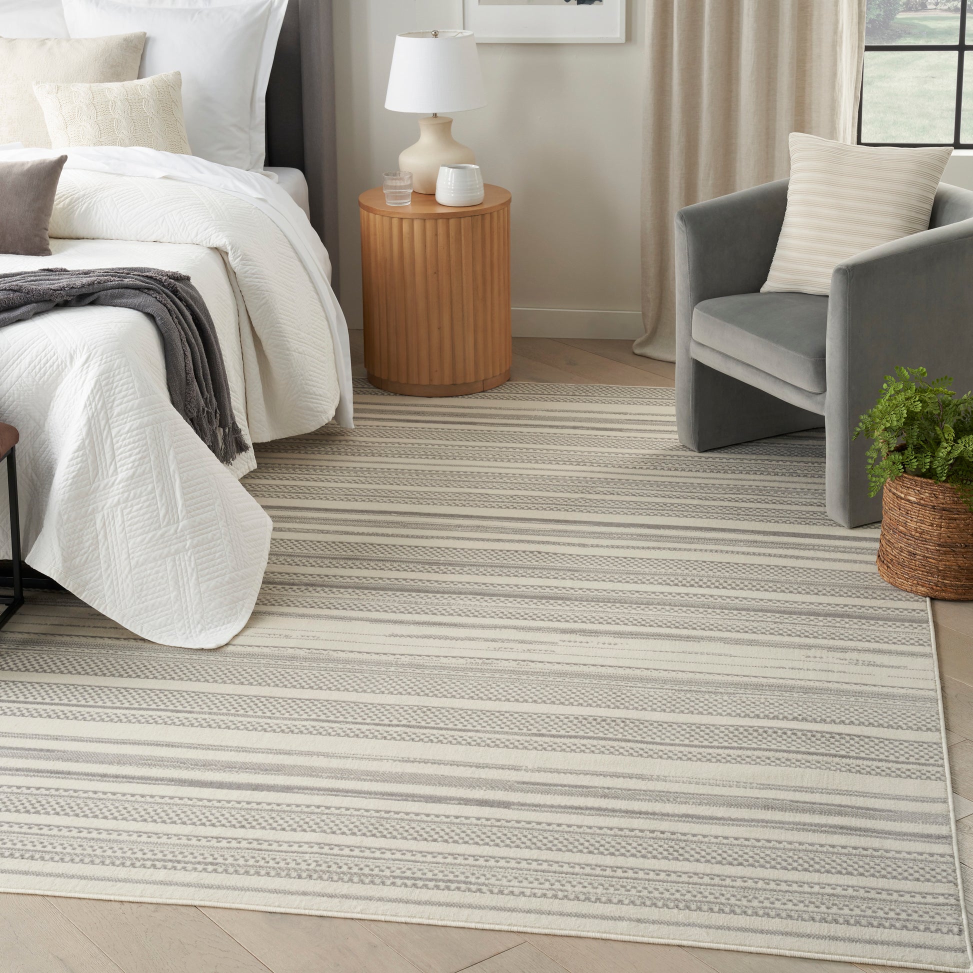 Neutral striped beige-gray rug beside a bed with round wooden nightstand, gray armchair and potted plant.