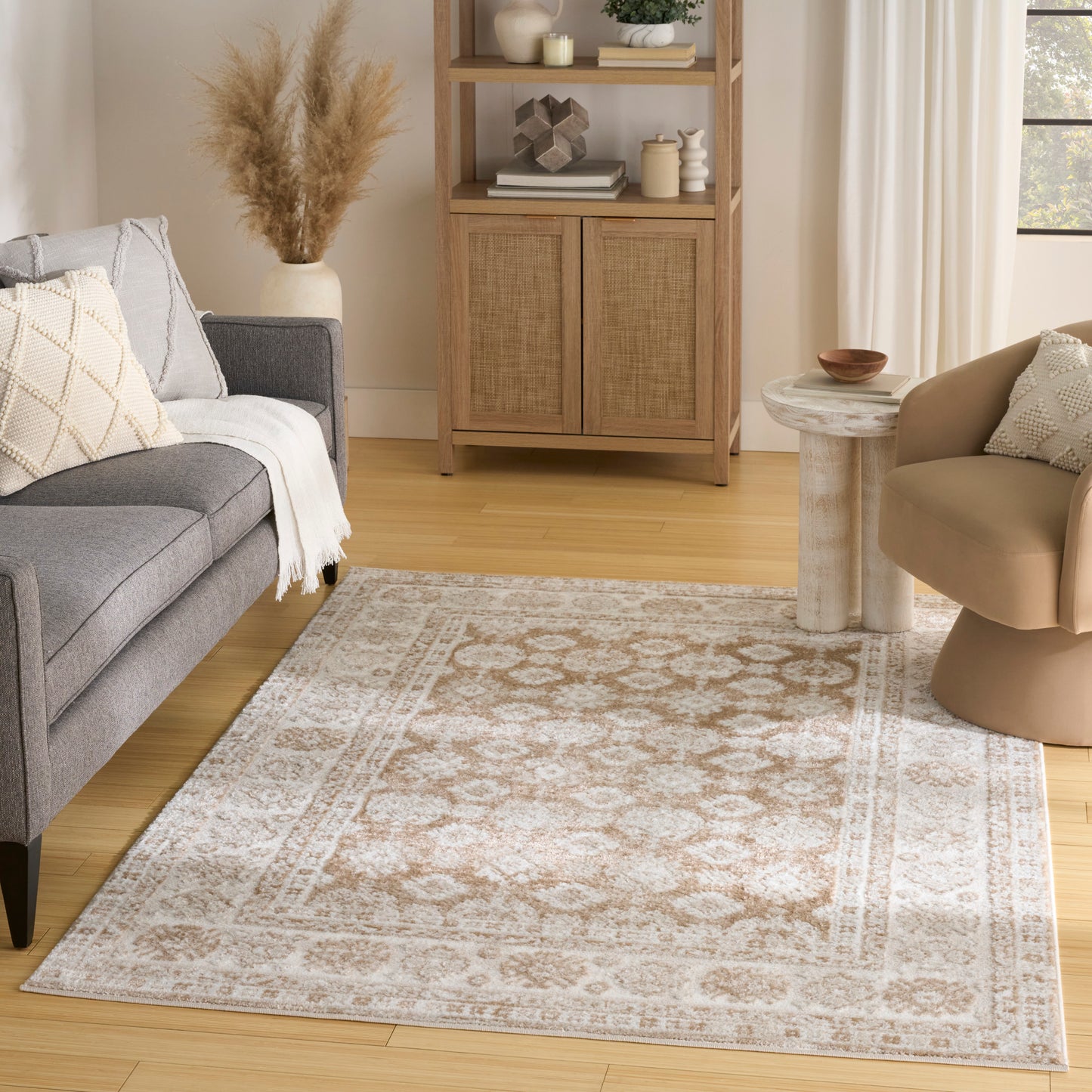 Cream patterned rug on a neutral living room floor with a gray sofa, tan accent chair, round marble side table, and a woven cabinet with decor and pampas grass.