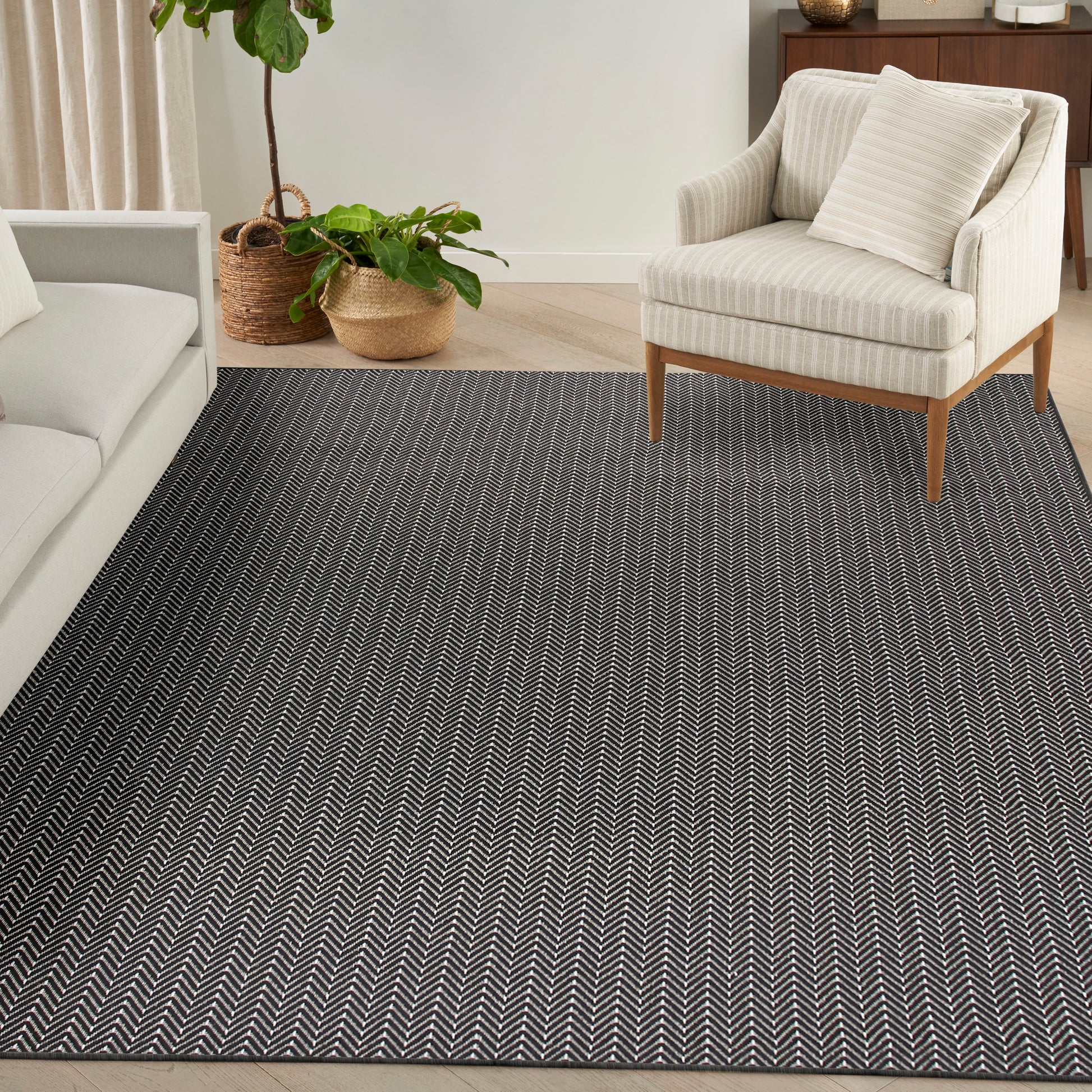 Dark gray chevron rug with white zigzag pattern in a modern living room; beige armchair, sofa, & woven plant baskets.
