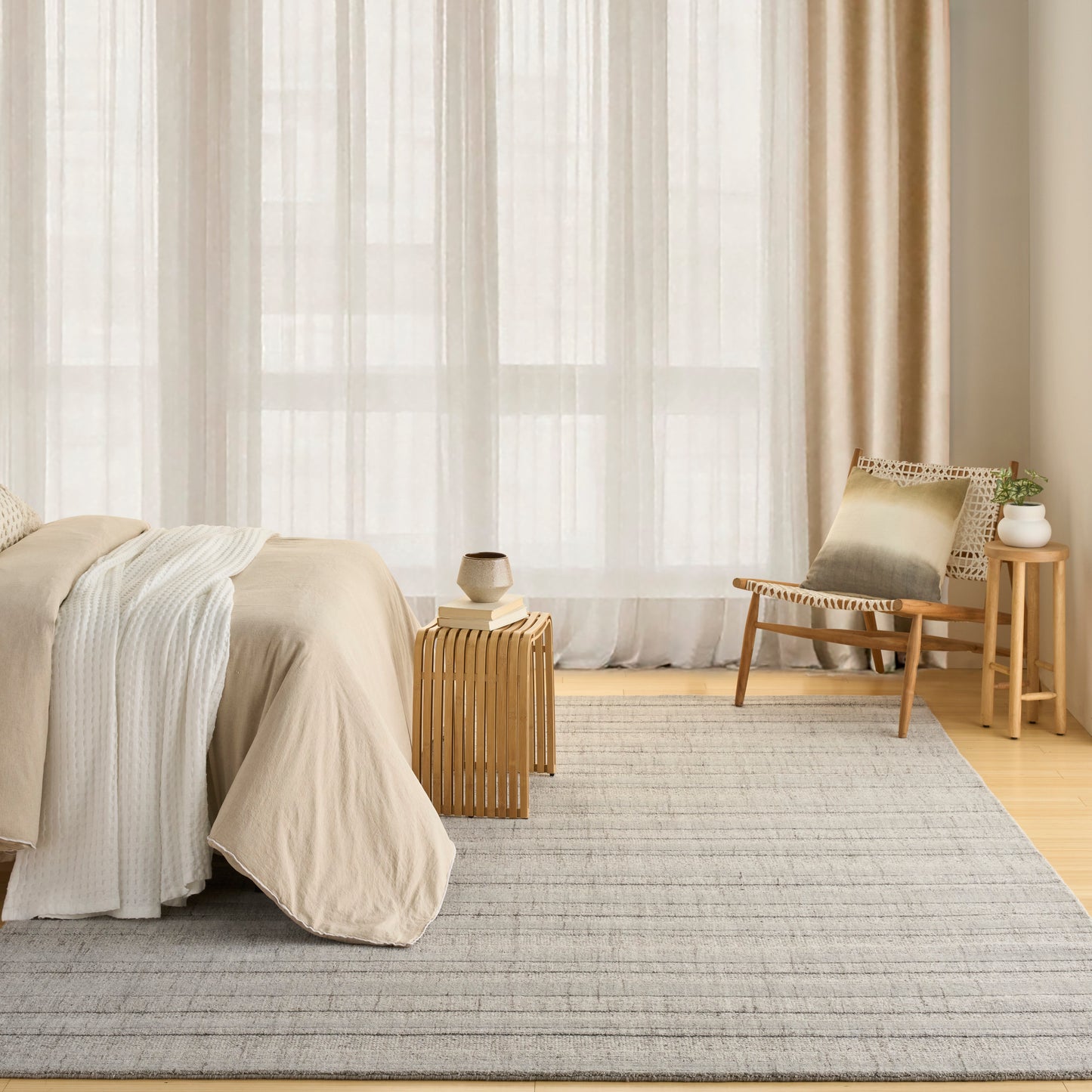 Gray rug in a neutral bedroom with beige bedding, light wood furniture, sheer white curtains & a woven lounge chair.