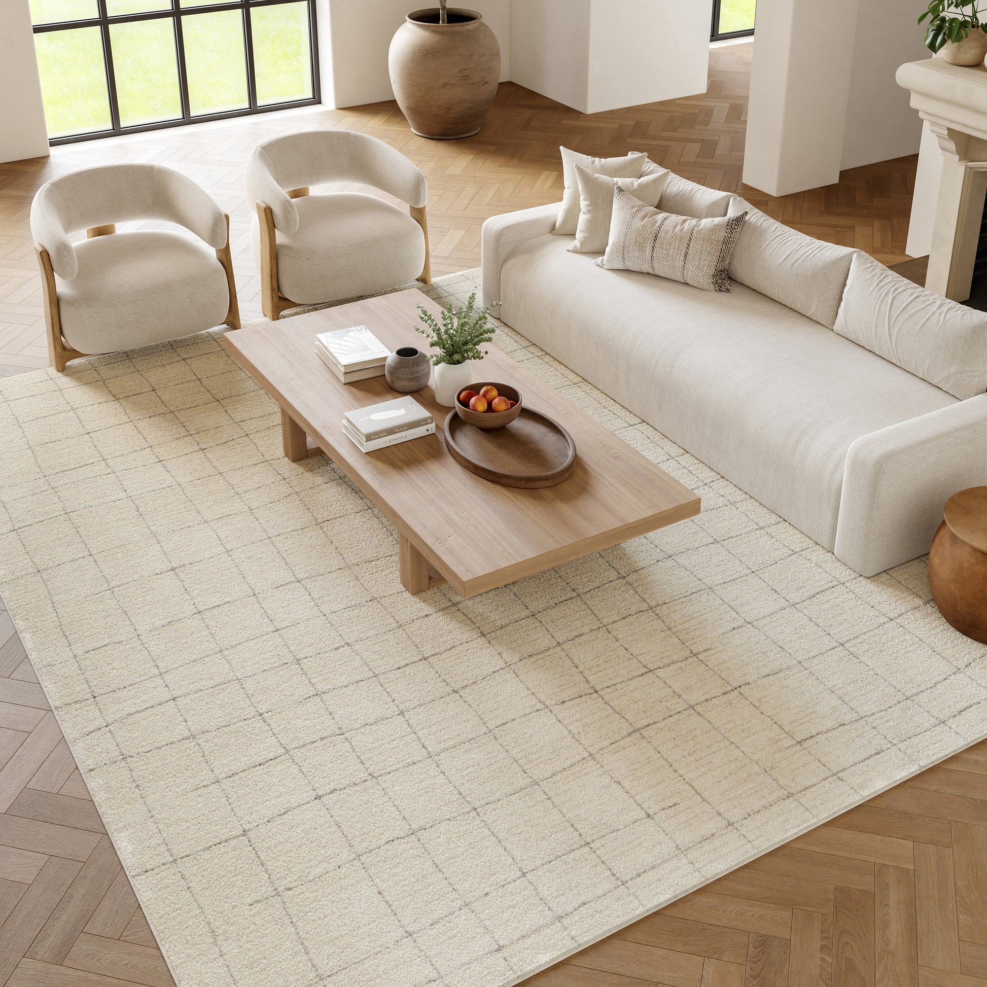 Modern living room with cream sofa, two curved beige armchairs, low wooden coffee table on a pale grid rug.