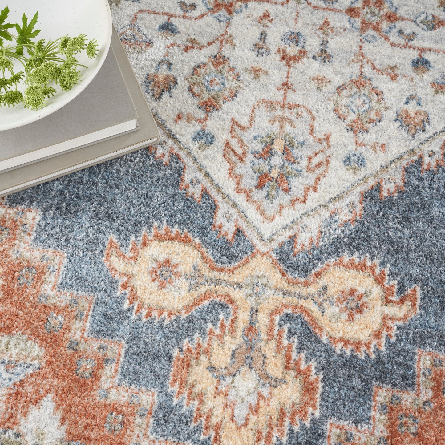 Traditional oriental area rug in blue, ivory, and rust with medallions; corner shows greens on a plate and stacked books.