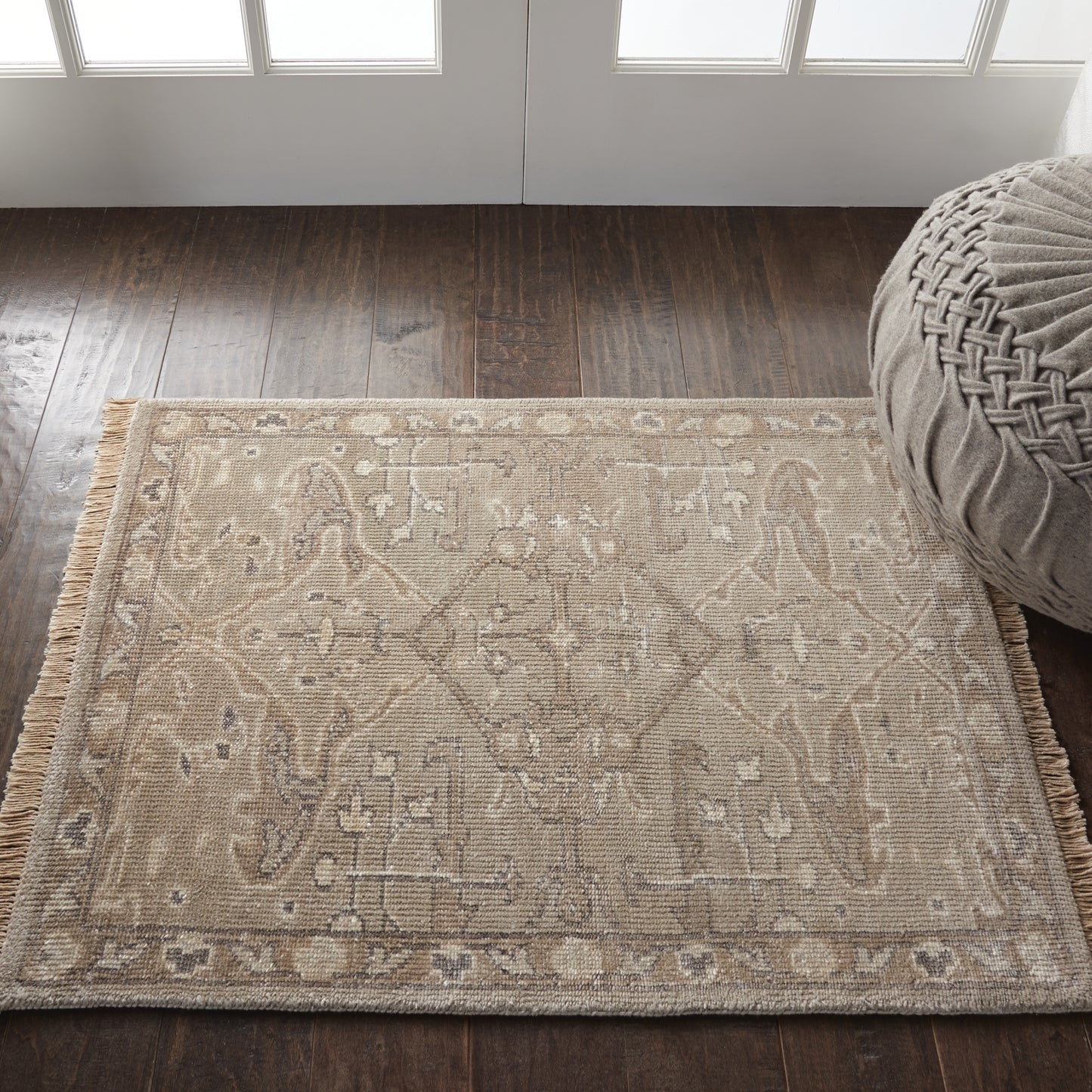 Beige flatweave rug with ivory geometric motif and fringe on a dark wood floor beside a round gray pouf.