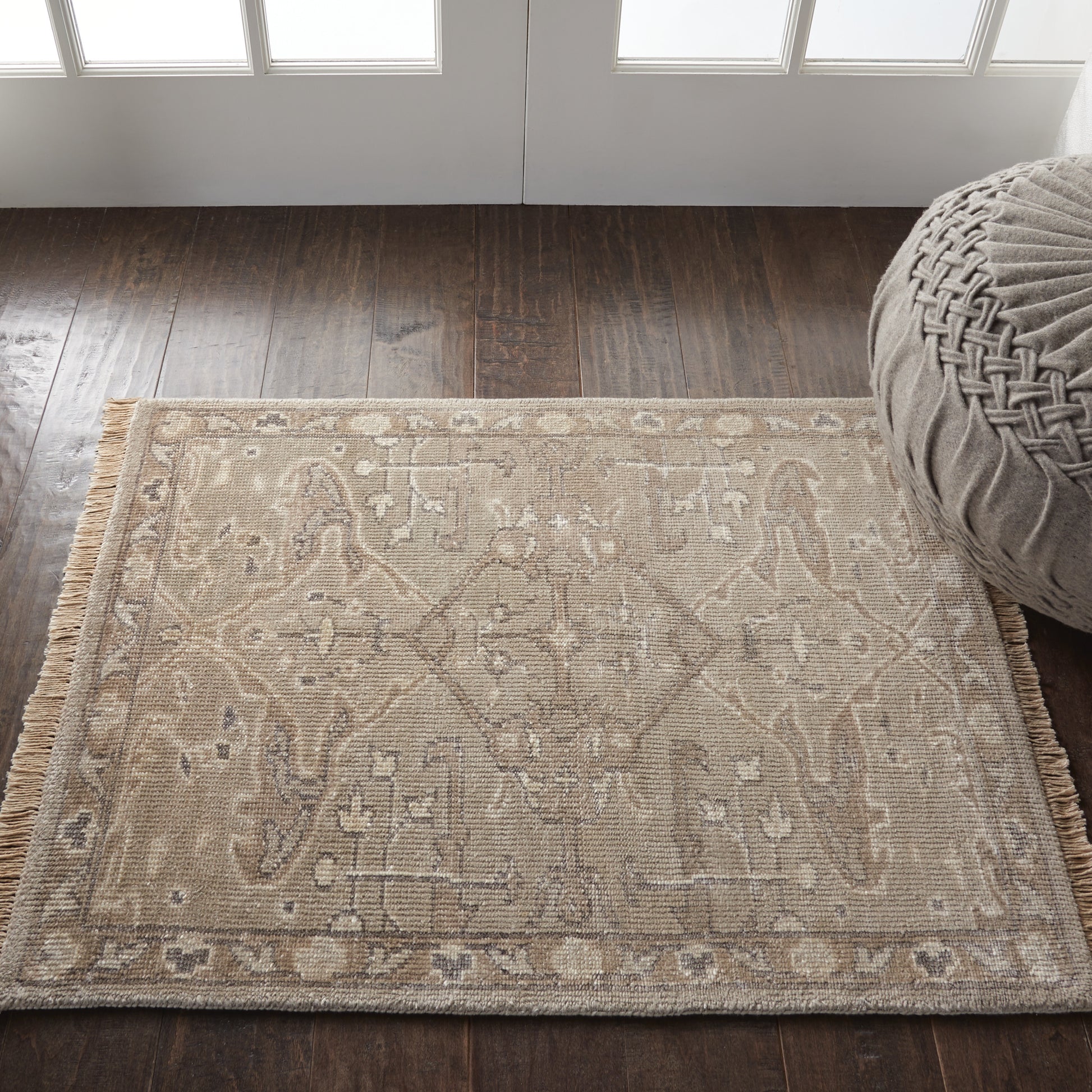 Beige flatweave rug with ivory geometric motif and fringe on a dark wood floor beside a round gray pouf.