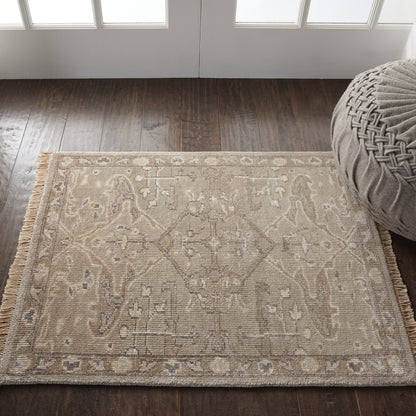 Beige flatweave rug with ivory geometric motif and fringe on a dark wood floor beside a round gray pouf.
