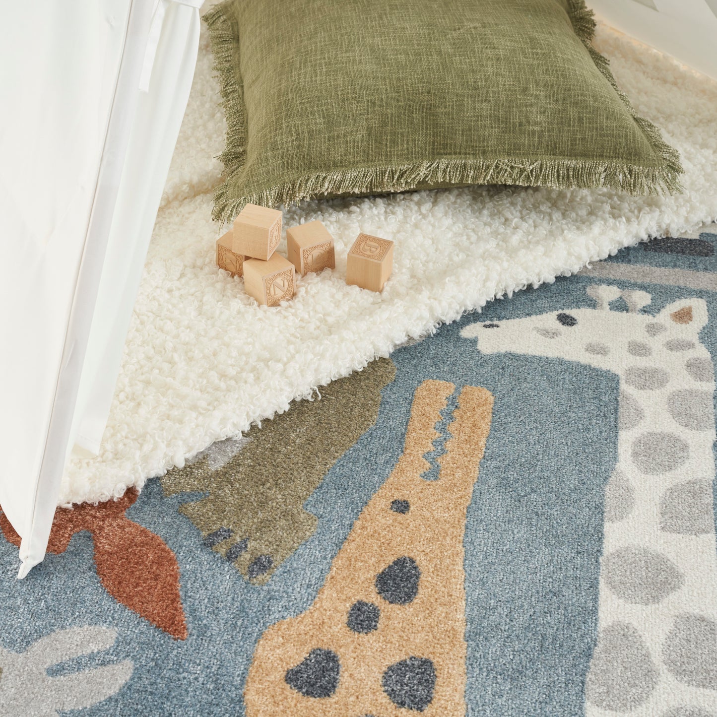 Green fringed pillow on a white shag rug with wooden alphabet blocks on a colorful animal-motif play rug; round 8' kids rug visible in the scene.