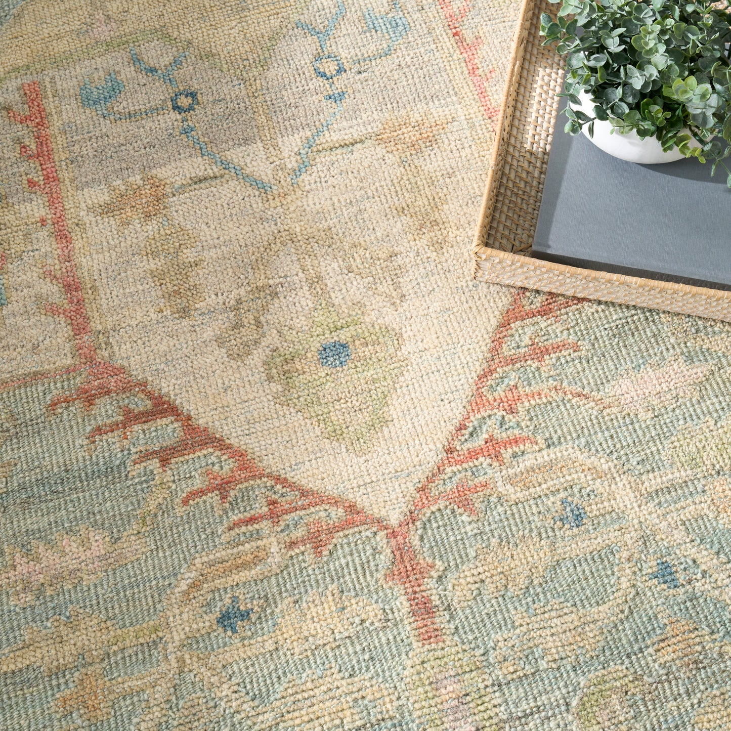 Pastel woven rug in beige with blue, pink & green motifs; textured pattern, corner view with a wicker tray & plant.