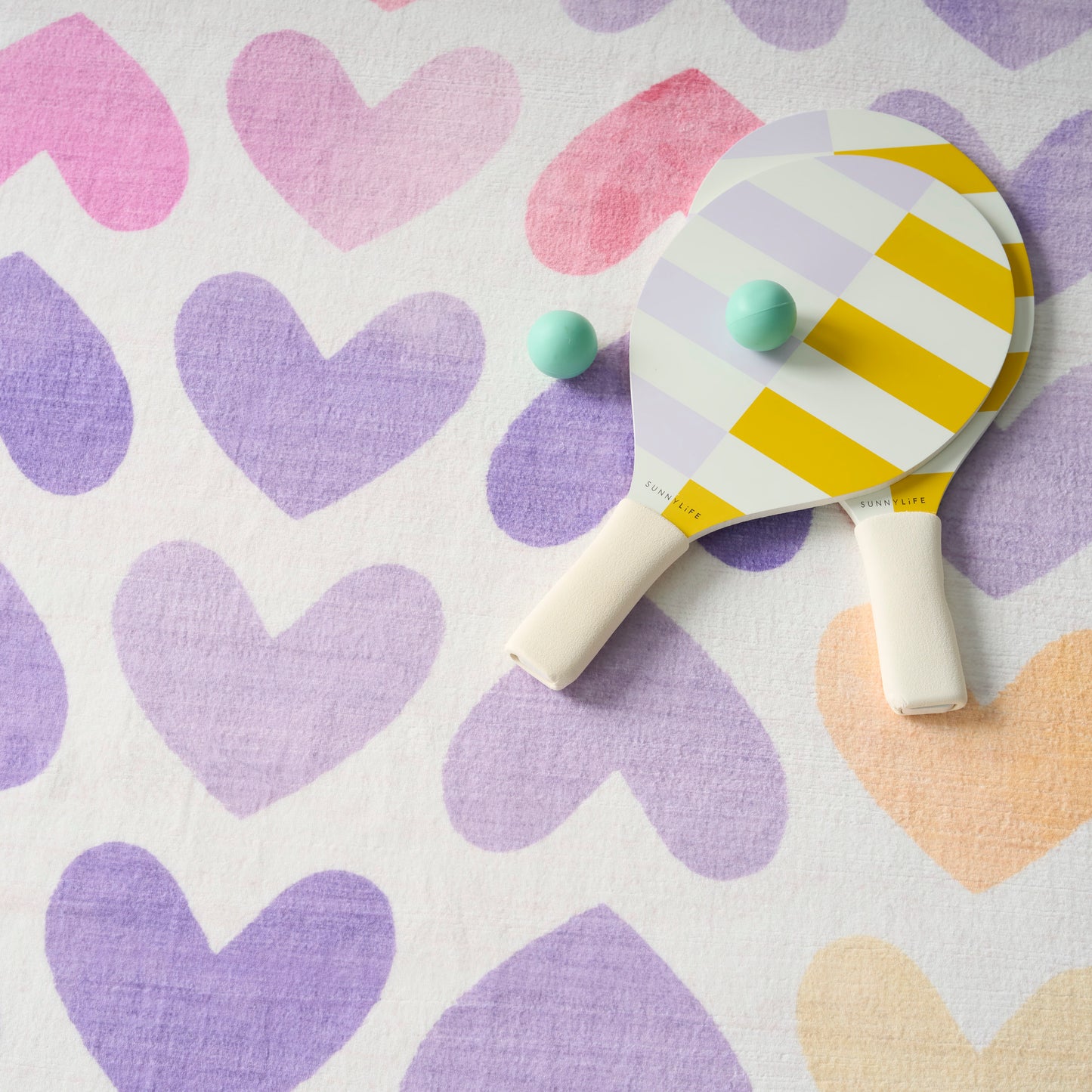 Pastel heart-pattern backdrop with a yellow-and-white striped ping pong paddle and two mint-green balls.