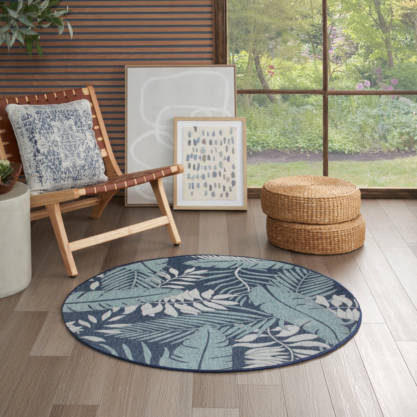 Round blue botanical rug on wood floor beside a wooden lounge chair, framed abstract art, woven ottoman, and garden view.
