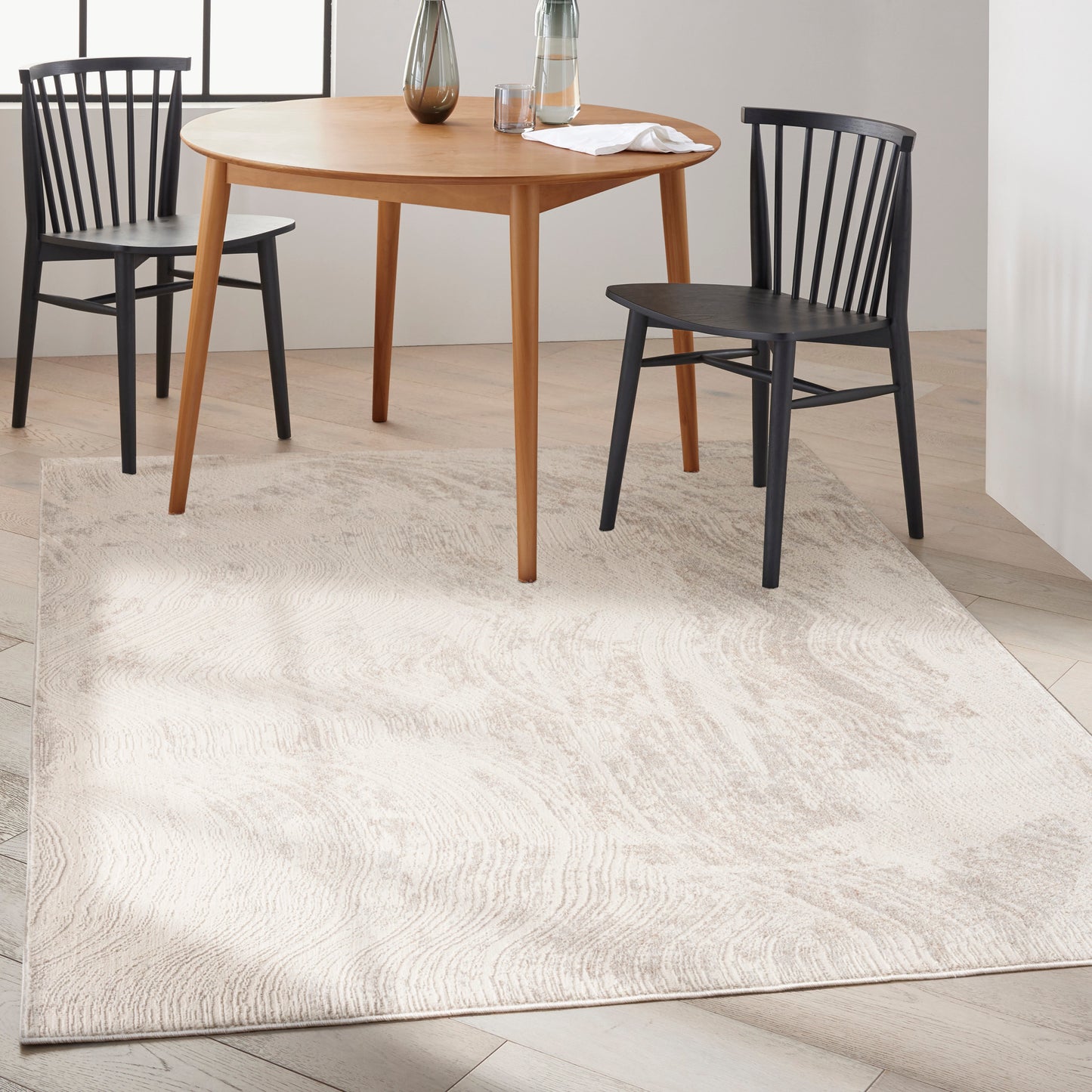 Round natural wood dining table with tapered legs and black spindle-back chairs; beige textured area rug.