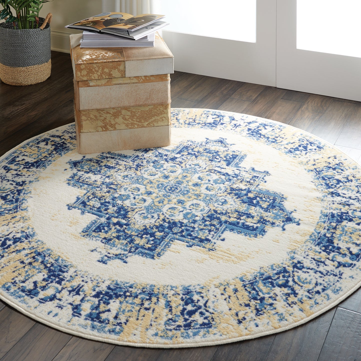 Round blue and cream oriental rug with tan accents on dark wood floor, beside stacked beige boxes and magazines.