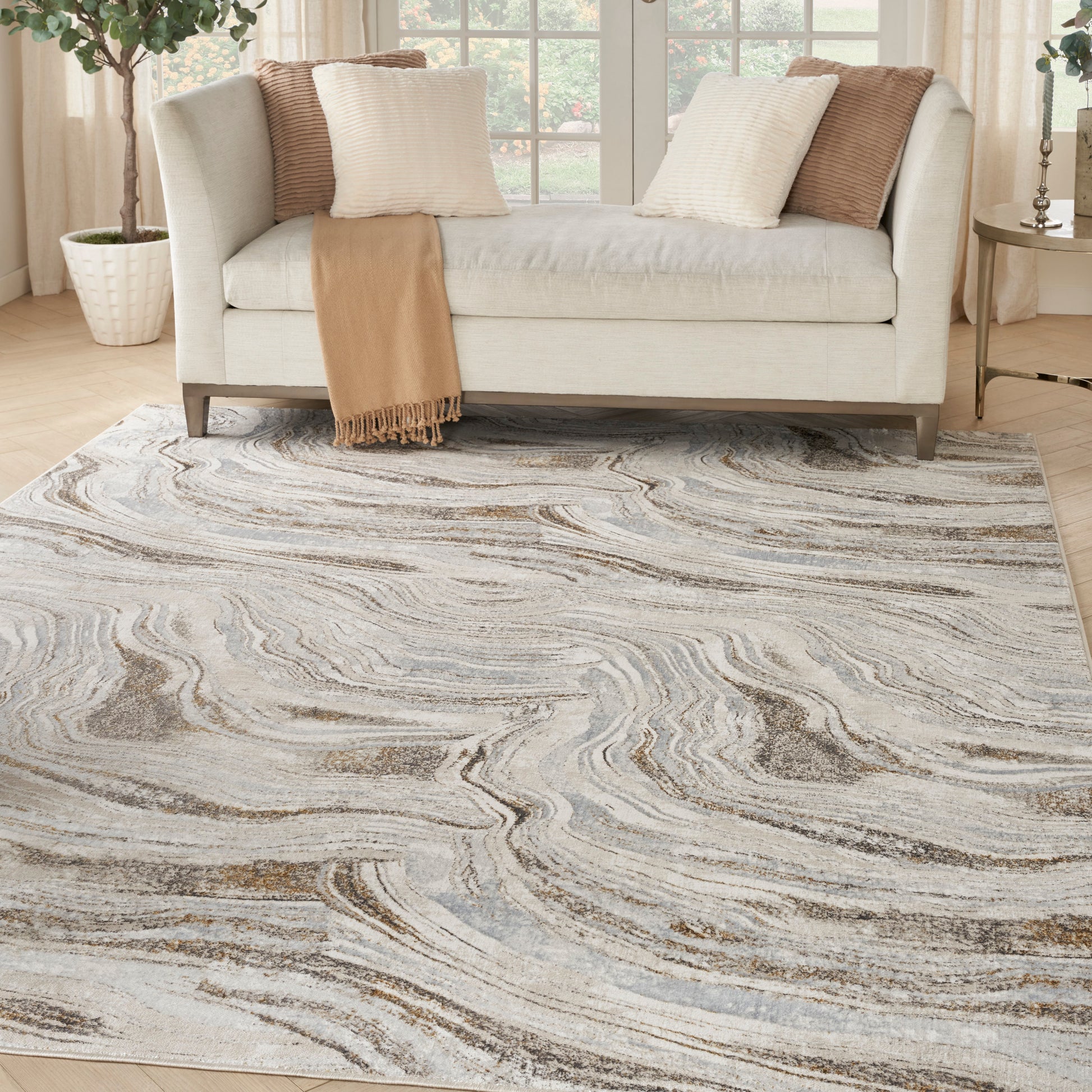Light gray sofa with cream and tan pillows, tan fringe throw, atop a swirling gray-beige area rug in a bright living room.