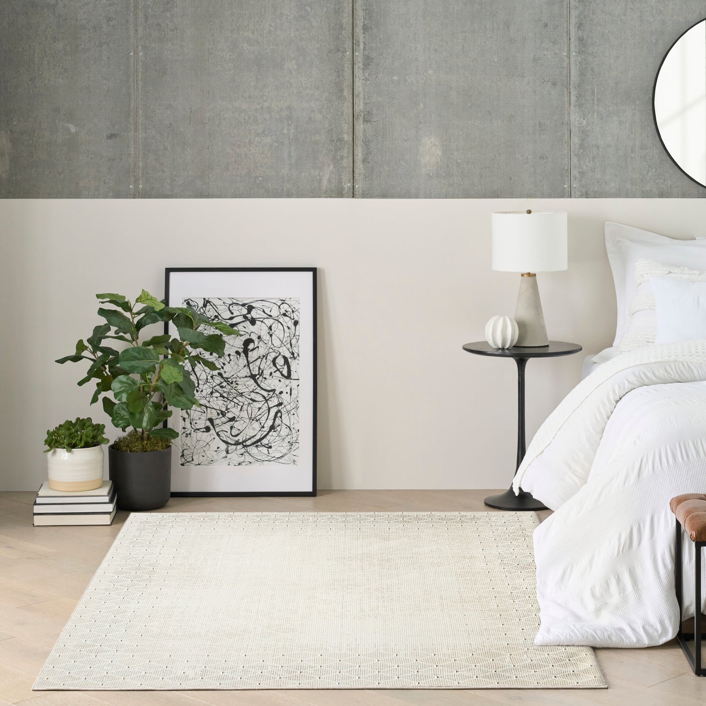 Minimalist bedroom with white bedding, round black nightstand, white lamp, concrete wall, framed abstract art, potted plant, beige rug.