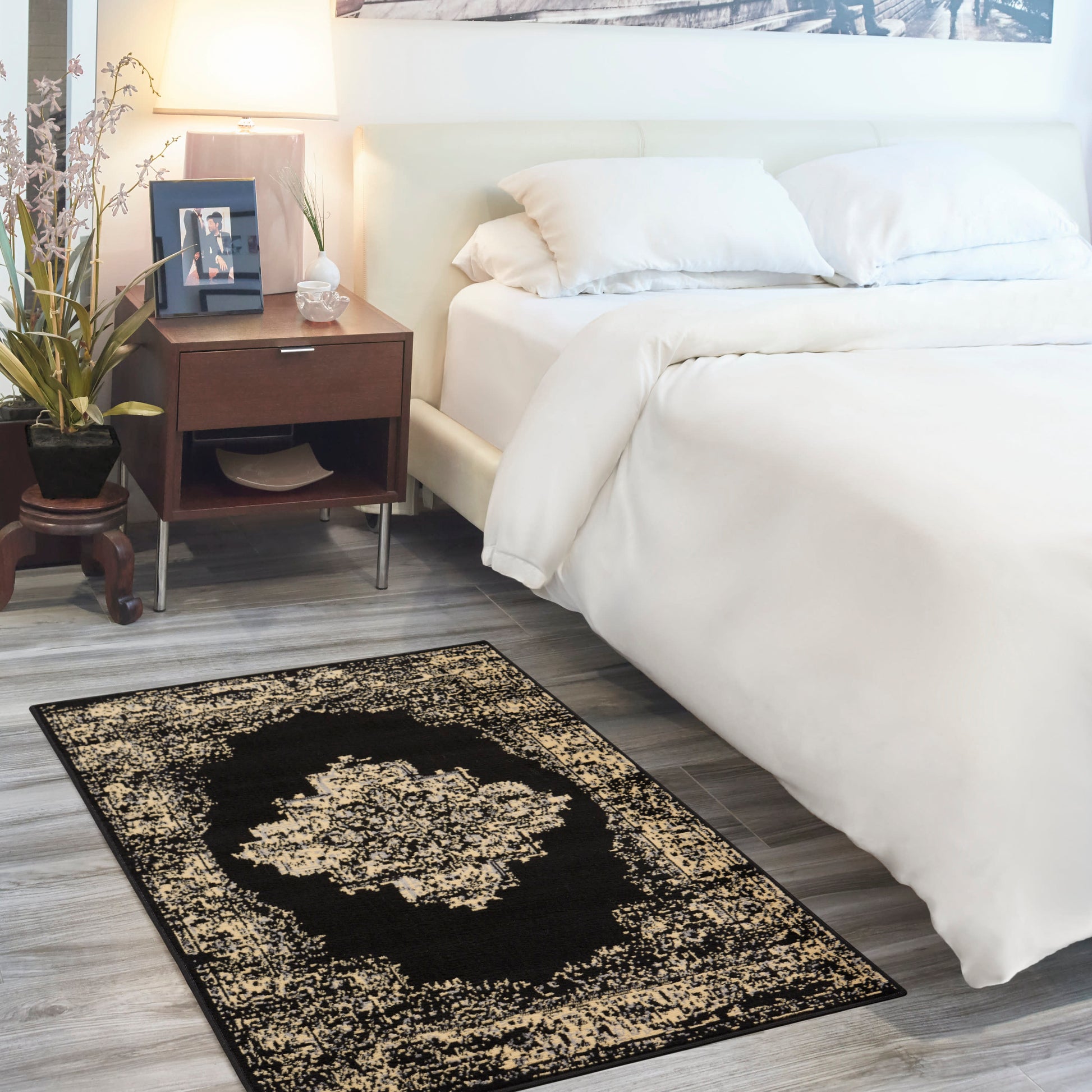 White bedding on an upholstered bed with dark wood nightstand, lamp, framed photo and plant; black-beige ornate rug on gray flooring.