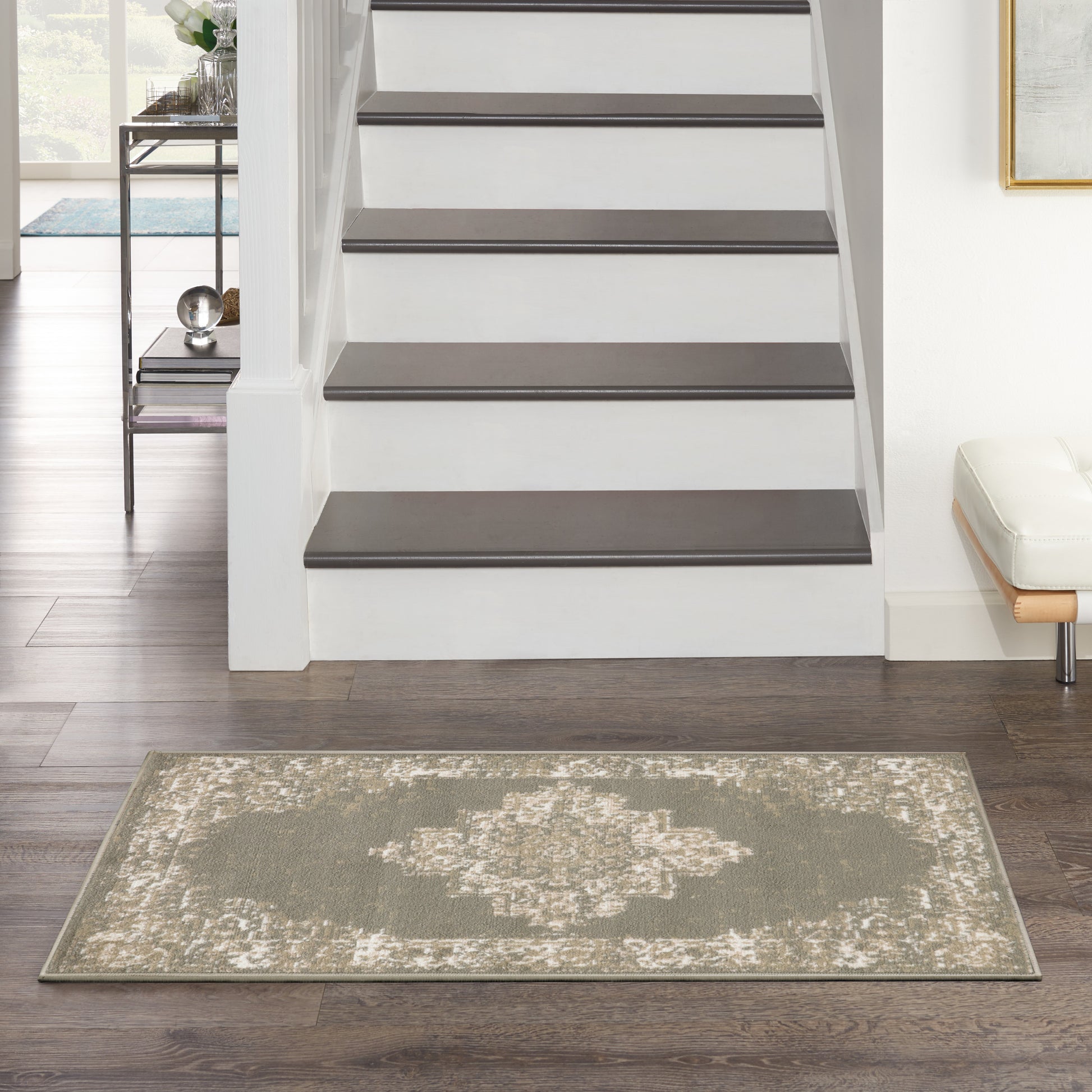 Rectangular olive-gray rug with cream geometric pattern on dark wood floor beside a white staircase in a bright living area.
