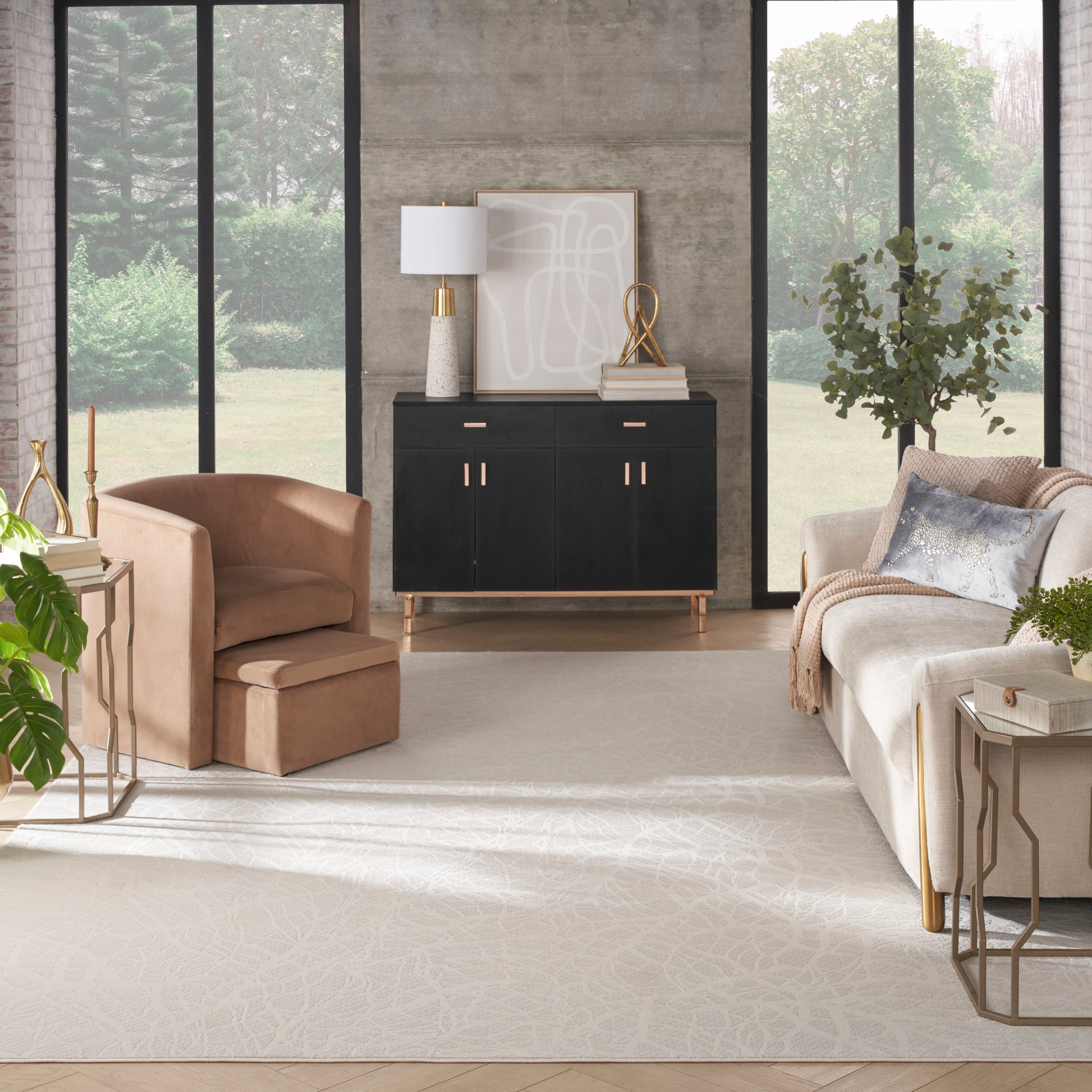 Modern living room with beige suede armchair, black cabinet with gold hardware, light rug, large windows, lush greenery.