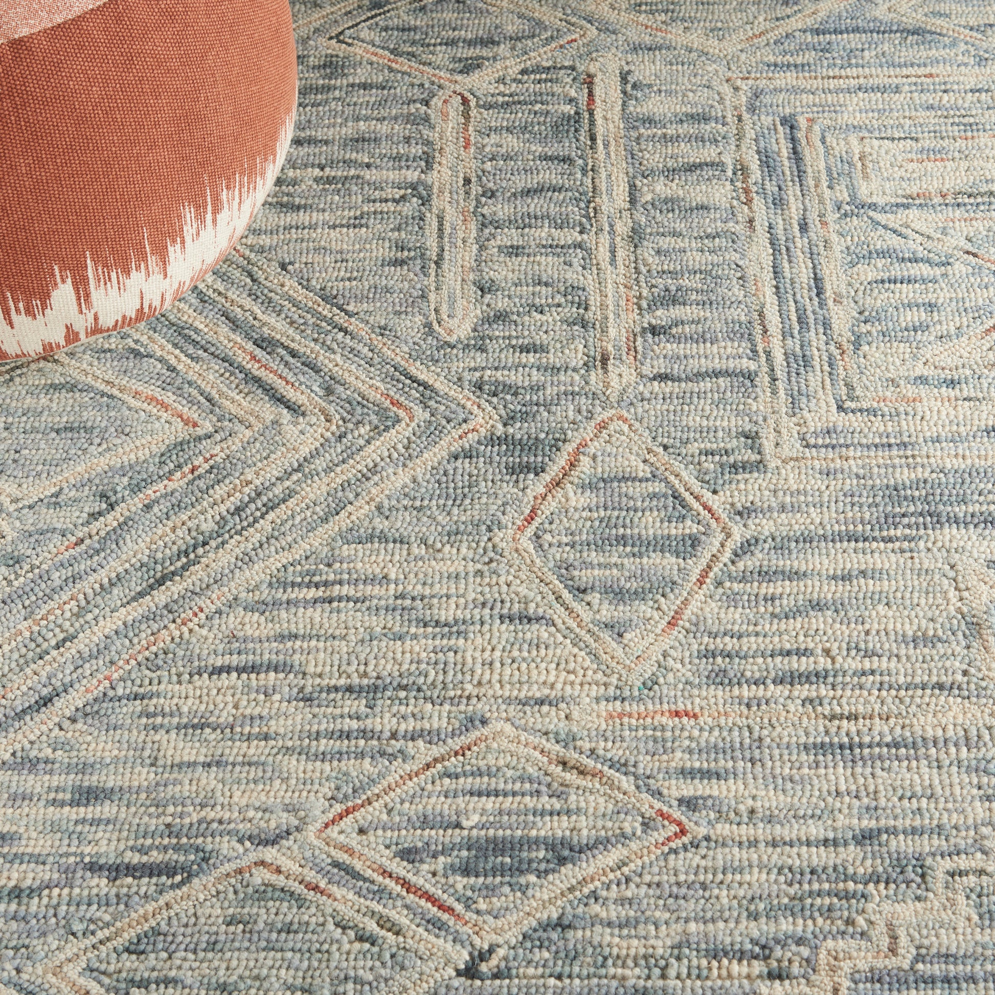Blue geometric woven rug with diamond motifs in rust outlines; round orange pouf peeks in top-left corner.