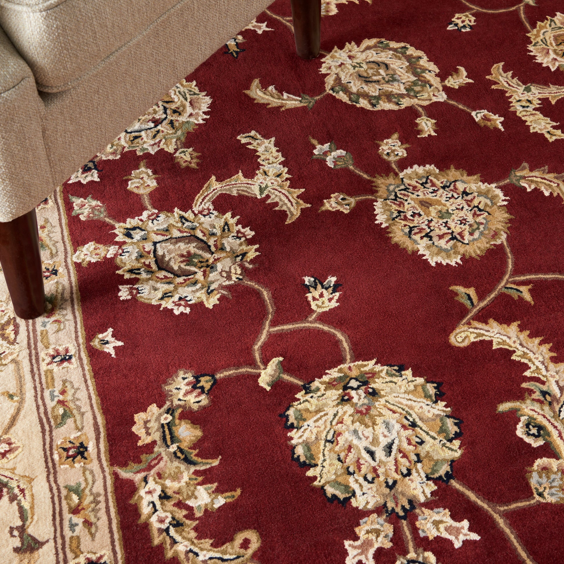 Burgundy oriental rug with large cream and gold floral motifs, bordered by a light pattern; wooden sofa leg in corner.