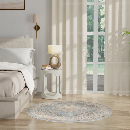 Neutral round rug in a cozy bedroom setting with white bedding, white nightstand, sheer curtains, and glass doors to a garden.