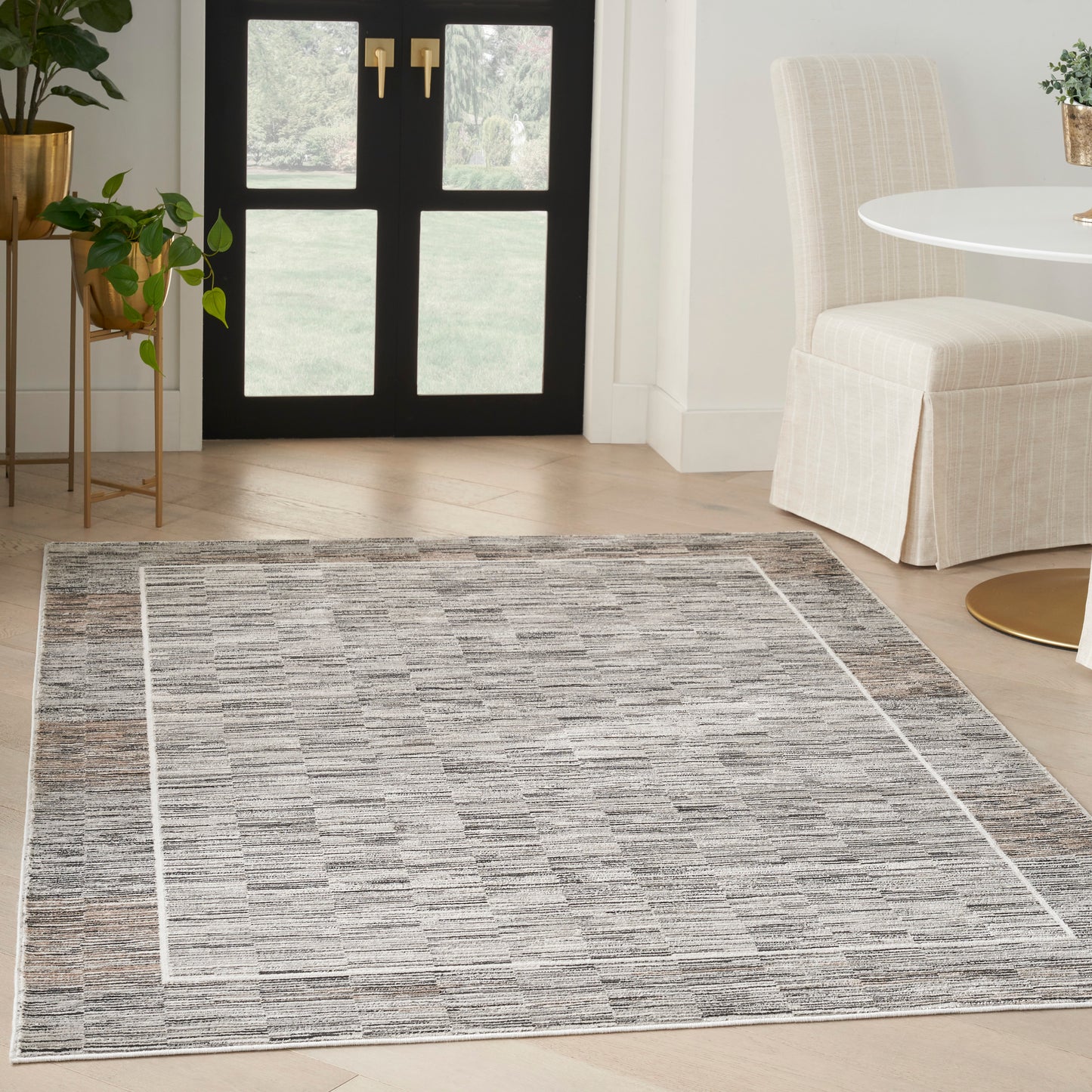 Grey textured area rug with white border in a light entryway, near black glass doors and beige upholstered chair.
