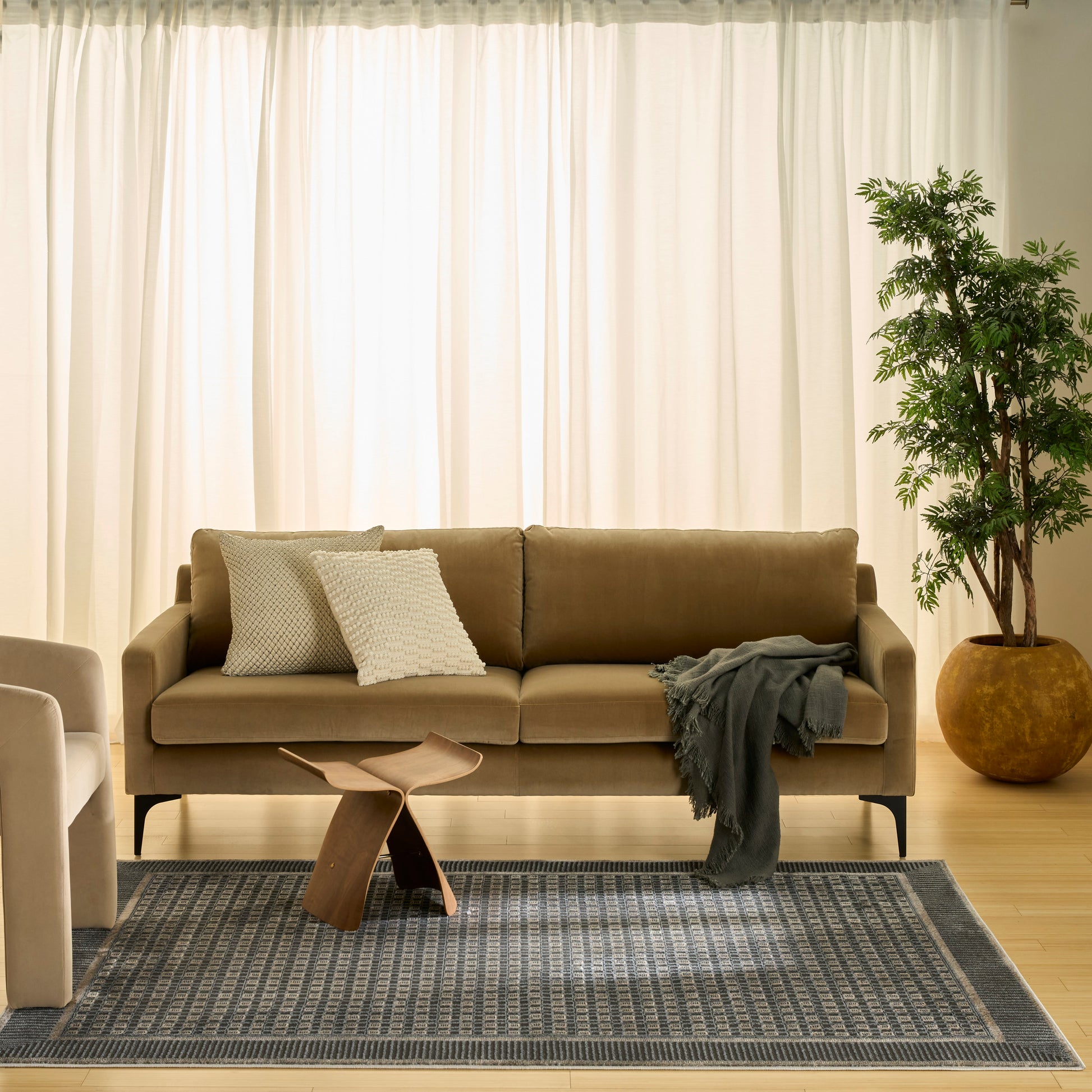 Beige fabric sofa with textured white pillows, gray throw, wooden sculptural coffee table, blue-gray rug, potted plant.