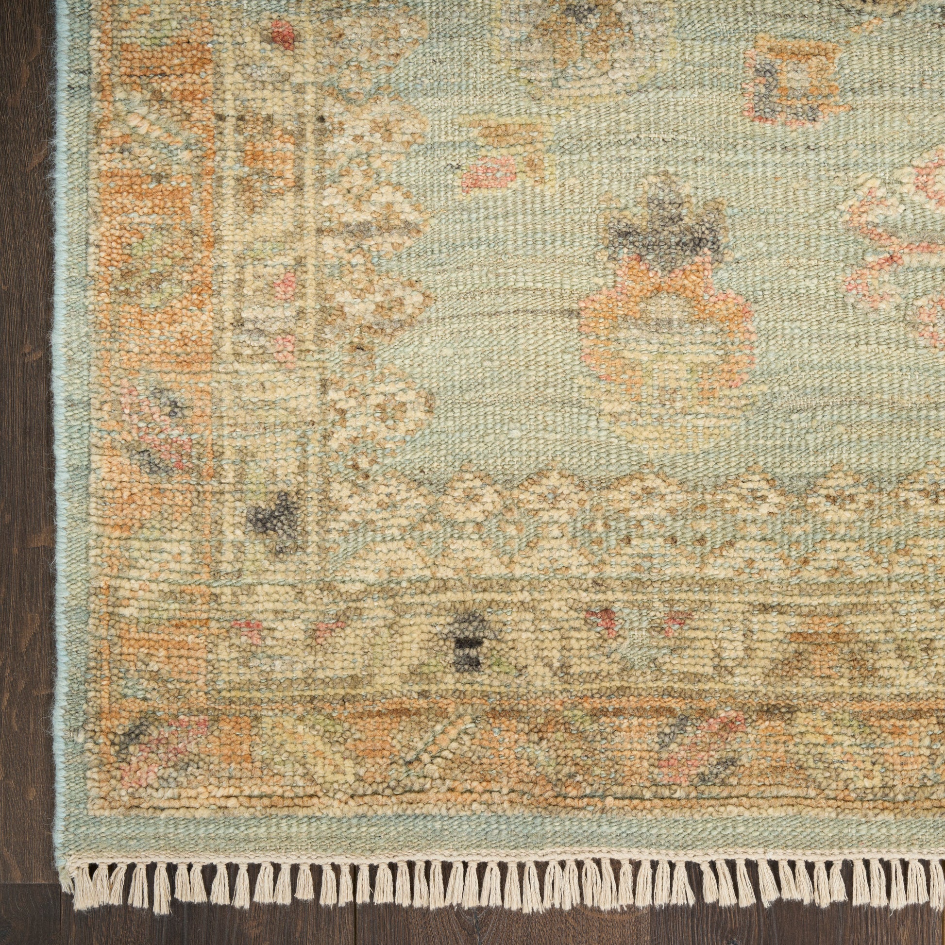 Pastel oriental rug with fringe, soft blues and beiges, subtle floral border, placed on dark wood floor.