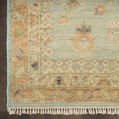 Pastel oriental rug with fringe, soft blues and beiges, subtle floral border, placed on dark wood floor.