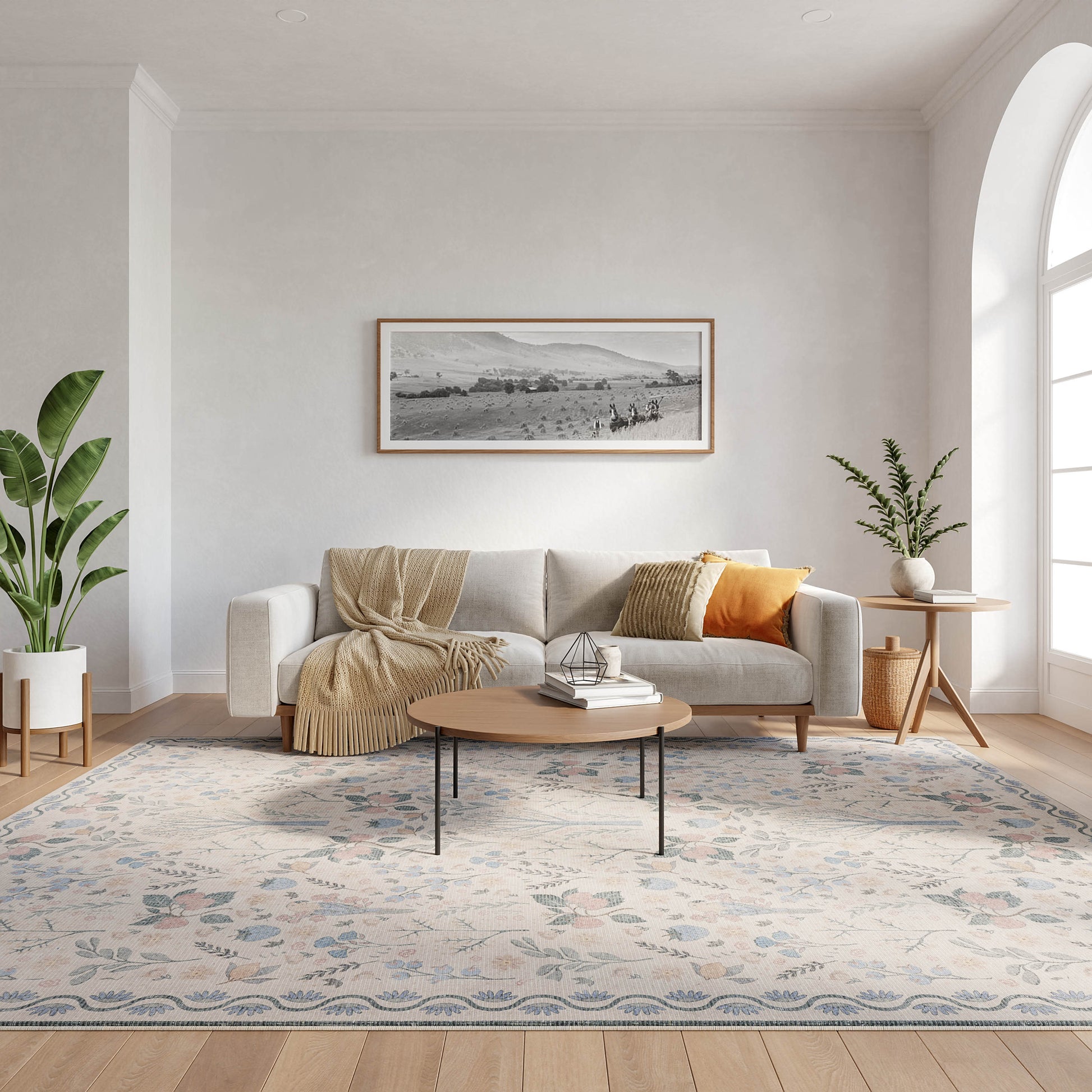 Bright living room with gray sofa, beige throw and orange cushion, round coffee table, patterned rug, green plants, window