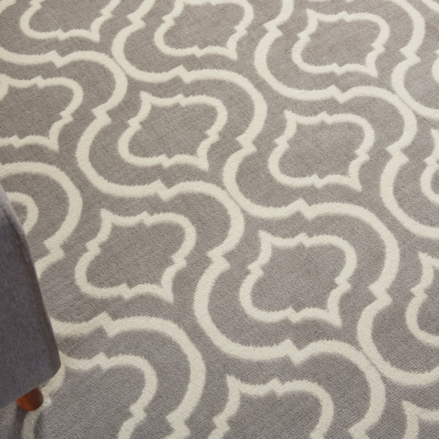 Gray area rug with white Moroccan trellis pattern; woven texture; corner of a sofa leg visible.