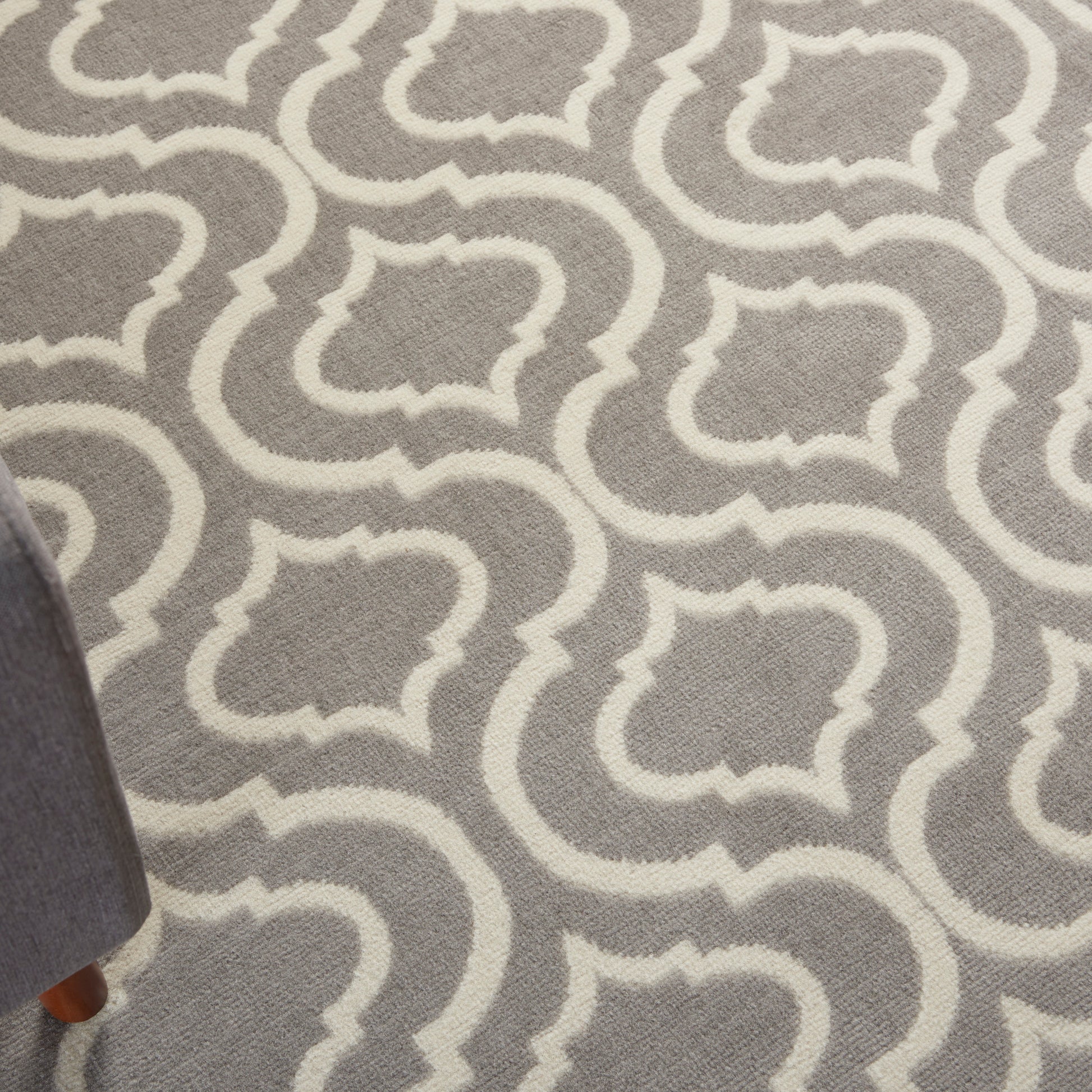 Gray area rug with white Moroccan trellis pattern; woven texture; corner of a sofa leg visible.
