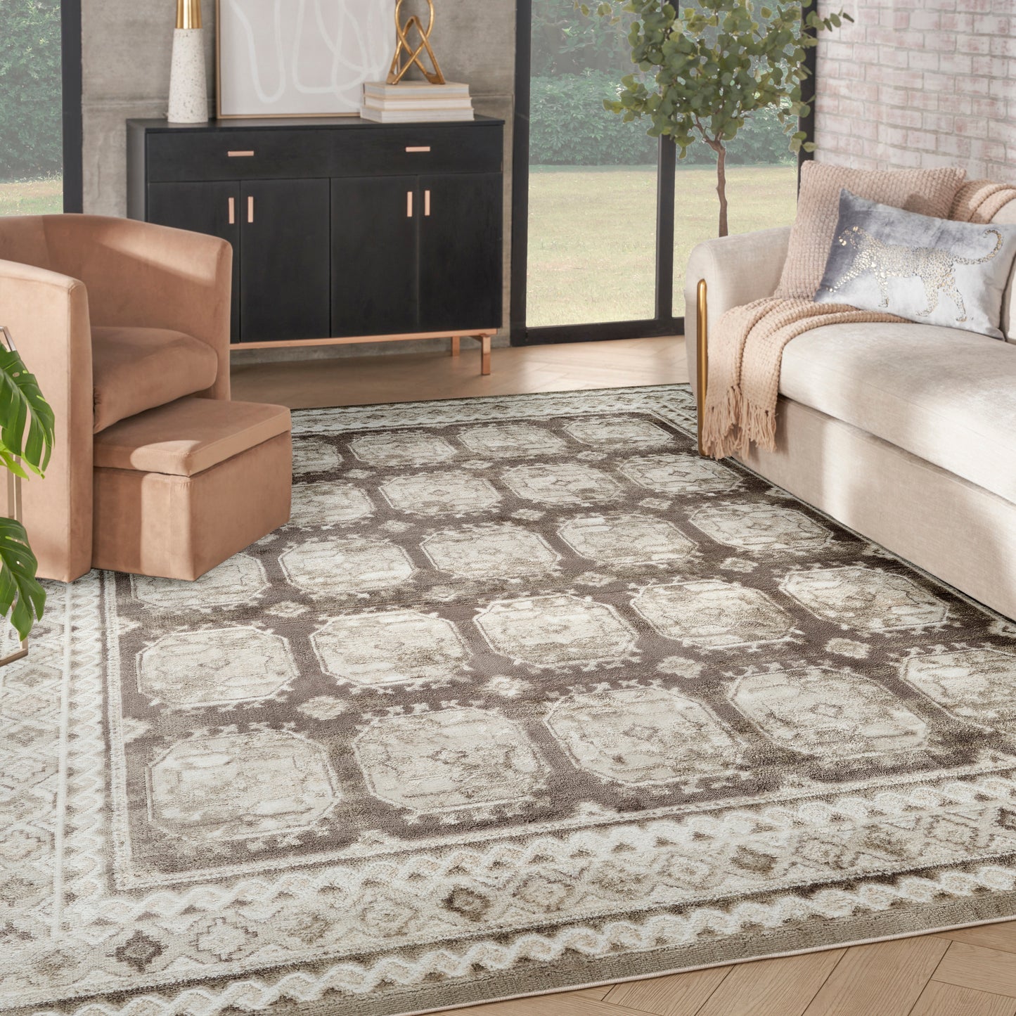 Beige-gray patterned area rug anchors a modern living room with tan chair, light sofa, black credenza, and big windows.