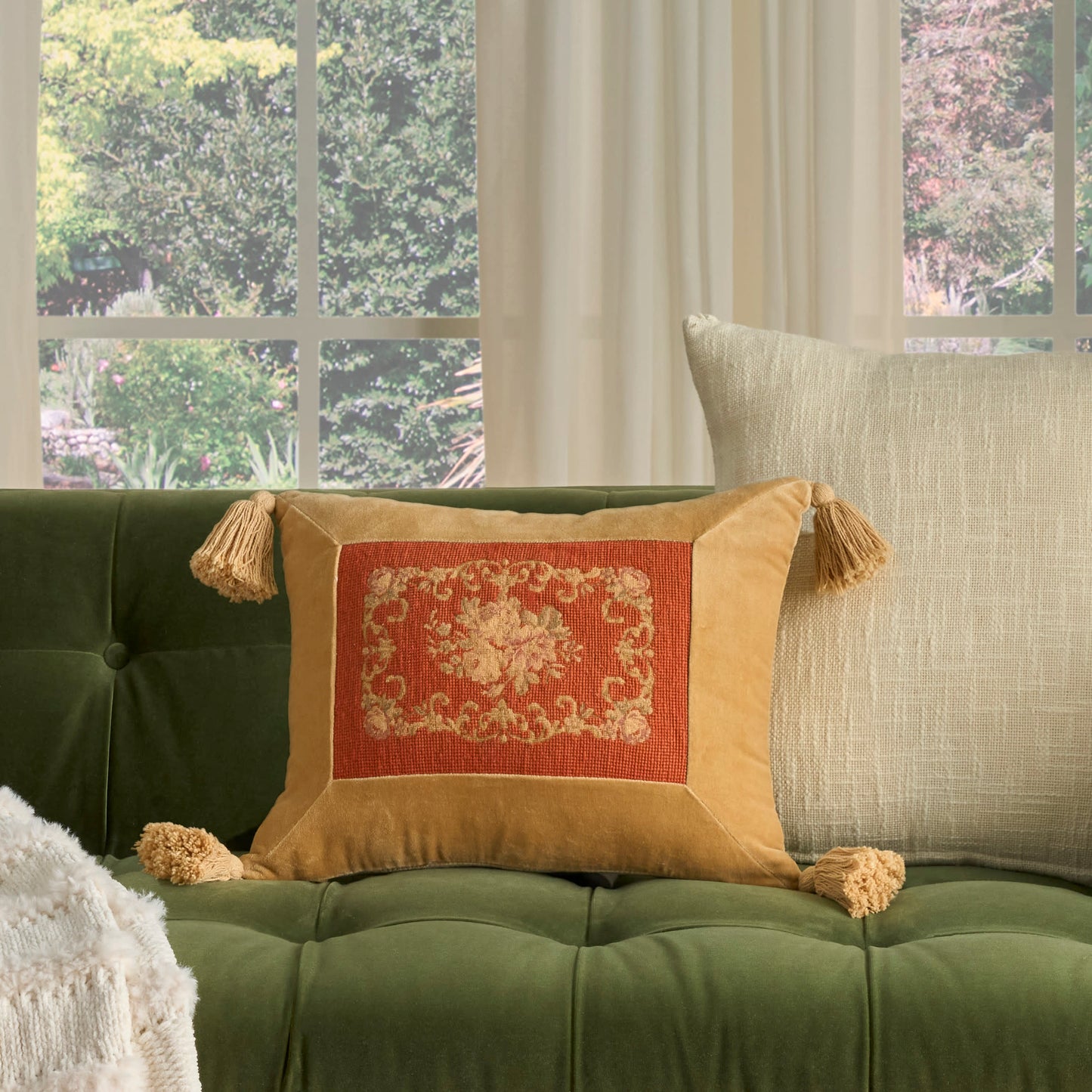 Green tufted sofa with an orange-gold decorative pillow featuring a floral center and tassels, beige cushion.