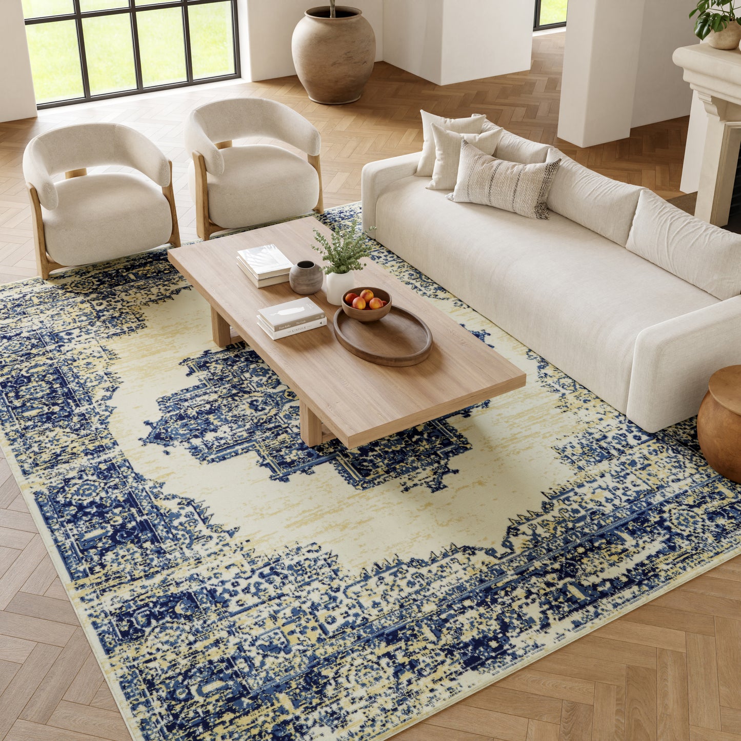 Beige sofa with two curved armchairs and a light wood coffee table on a blue-yellow patterned rug in a bright living room.