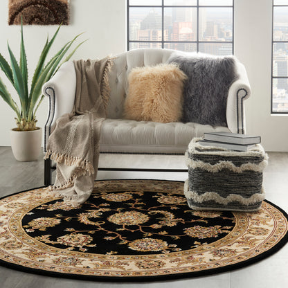 White tufted sofa with beige and gray pillows and throws, round black-gold ornate rug, potted plant, book-stacked ottoman.